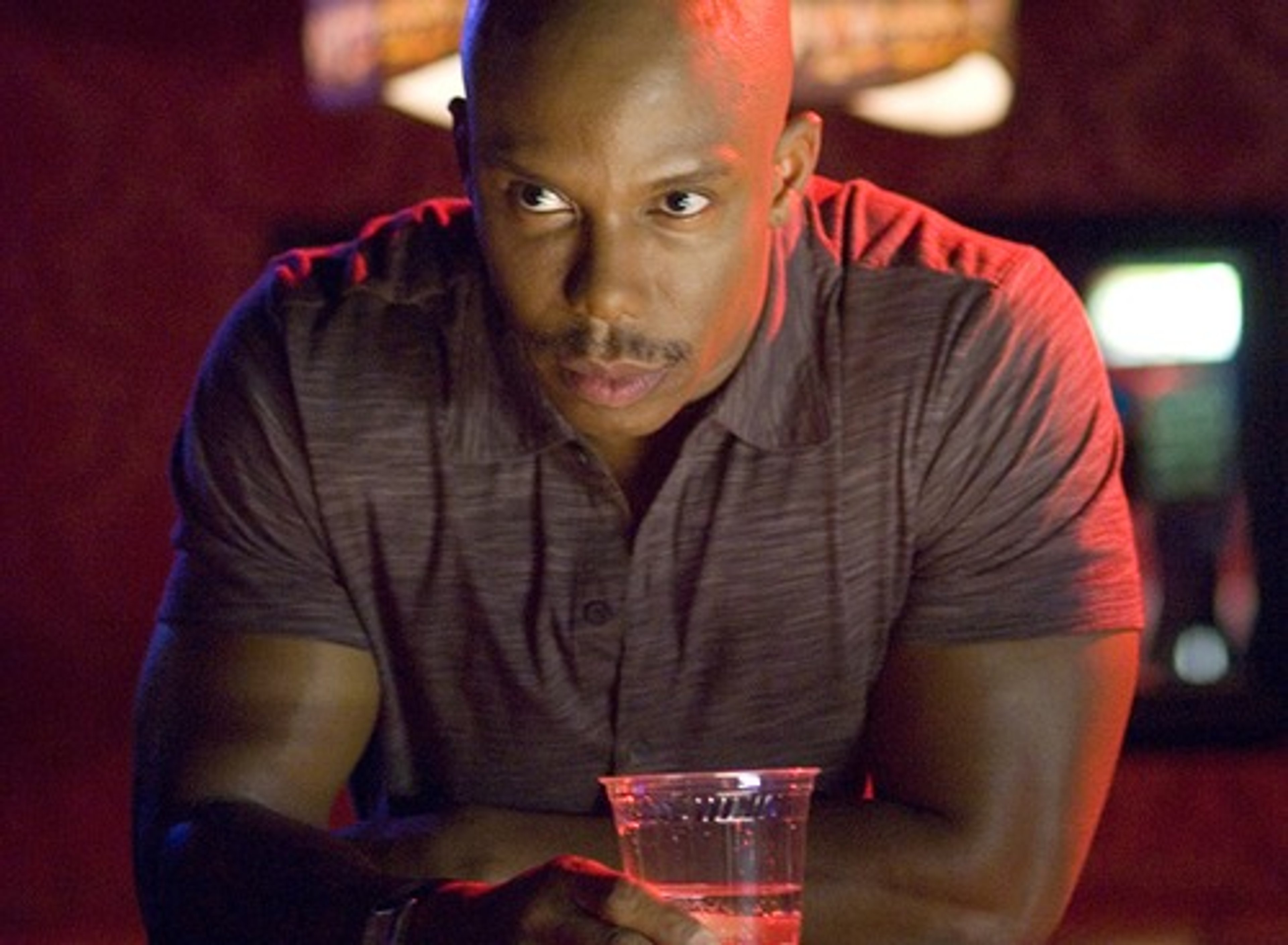 A close-up portrait of a serious-looking middle-aged Black man with a shaved head and muscular build.