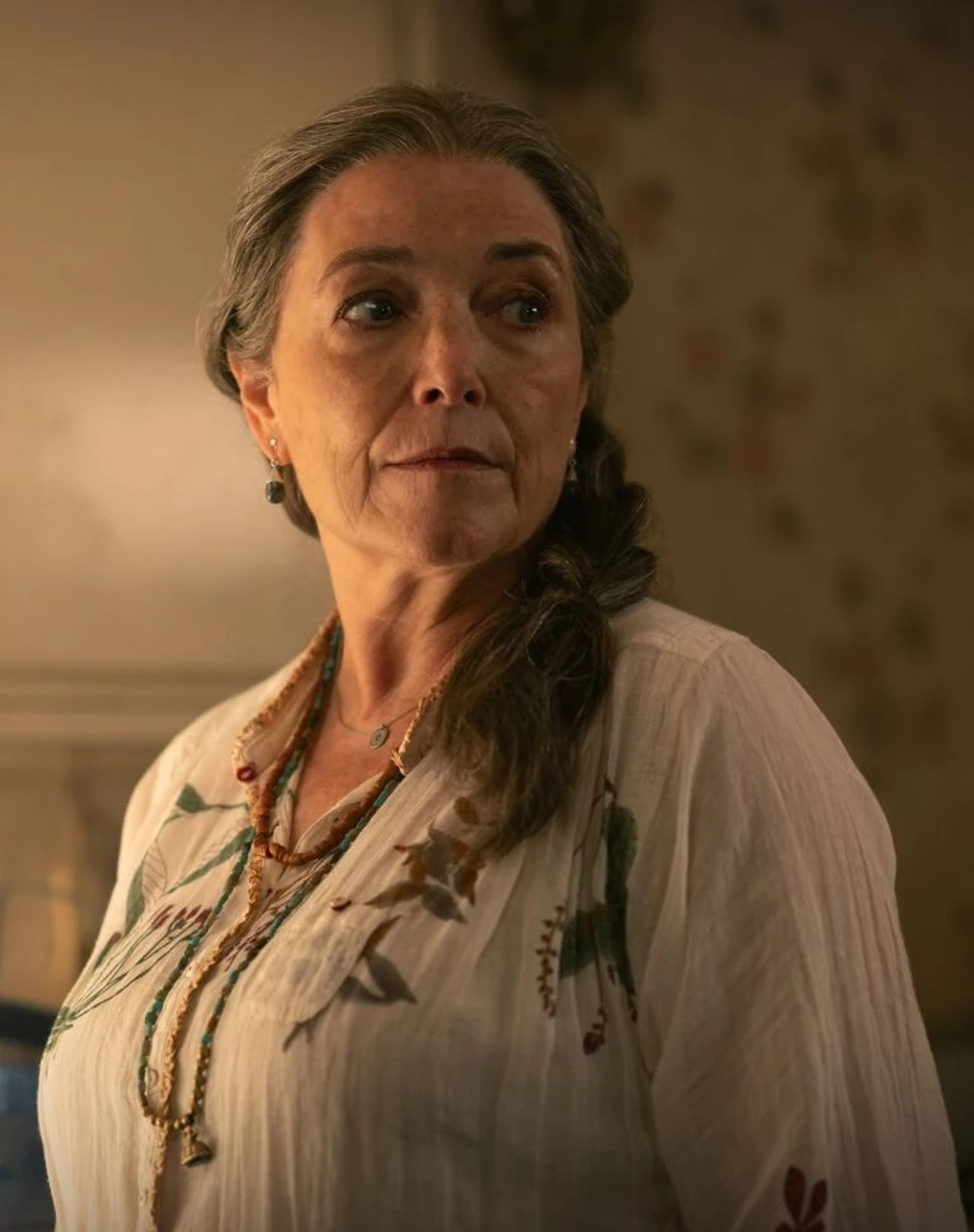 An older woman with gray hair in braids, wearing a white blouse and having a serious expression