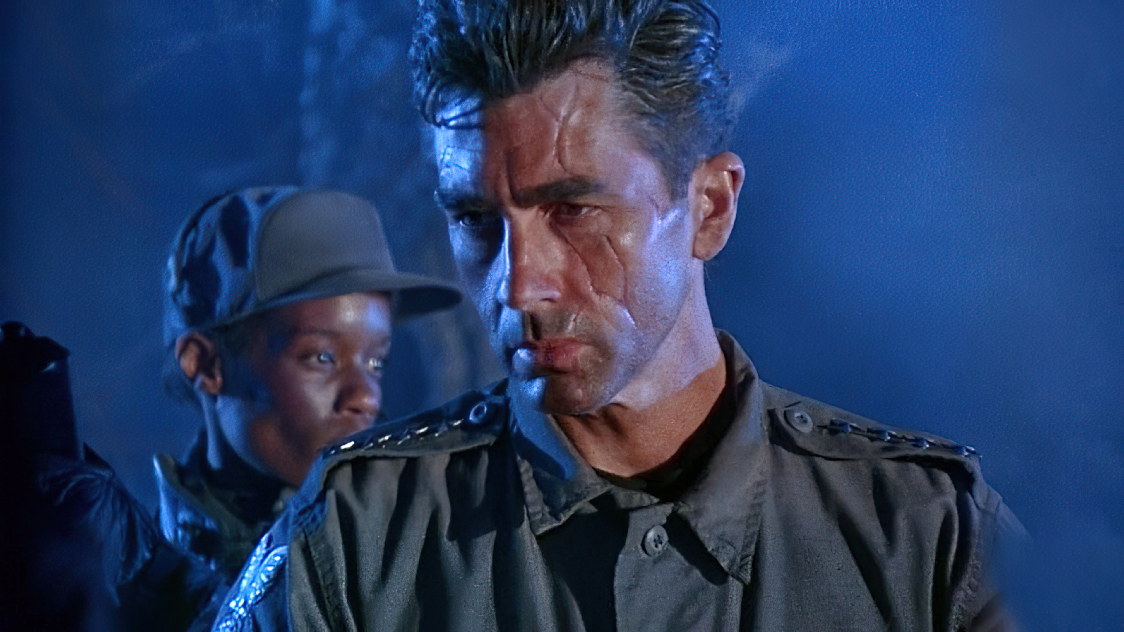 A man with short dark hair wearing a military jacket in a dimly lit industrial setting