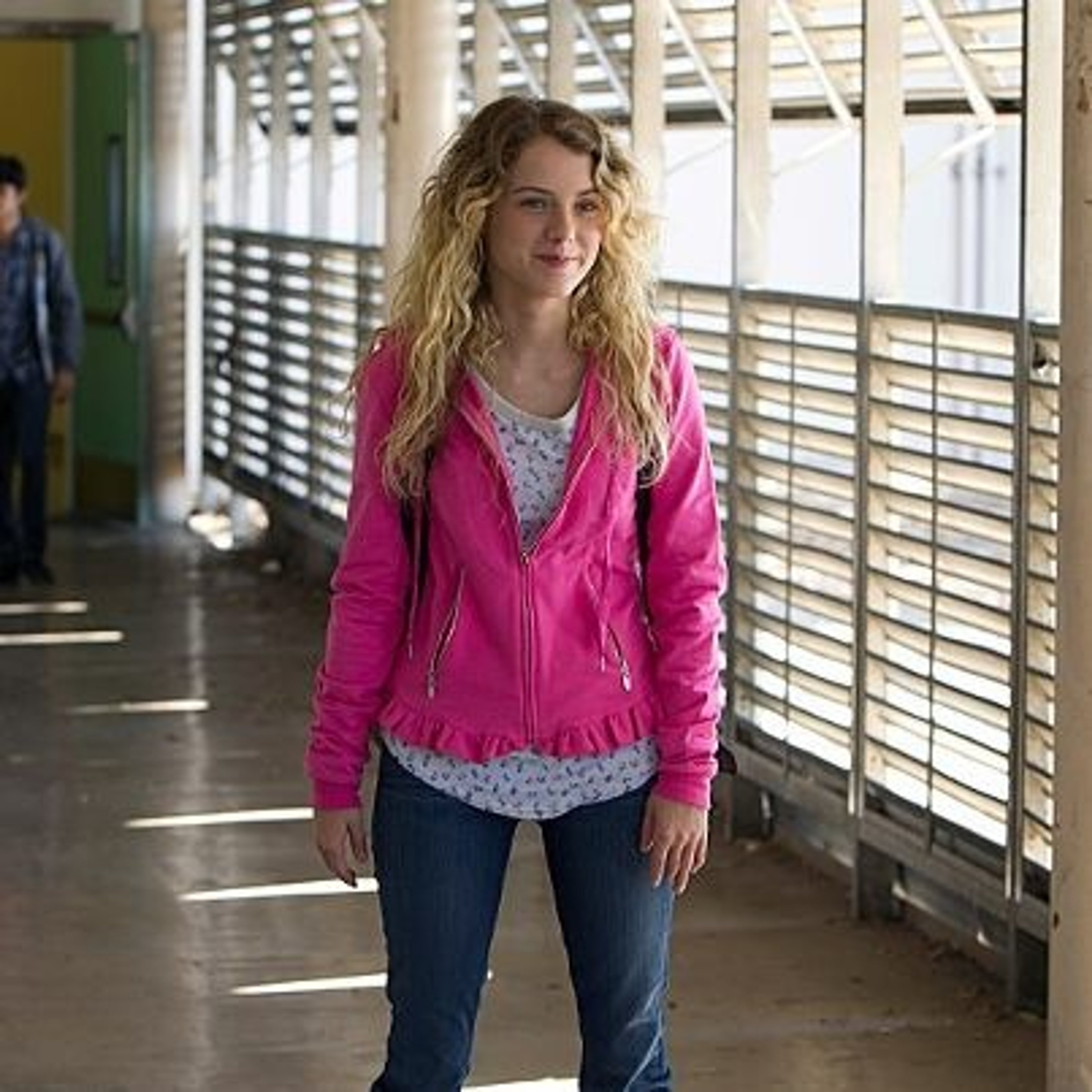 A young woman with curly blonde hair wearing a pink jacket standing in a school hallway