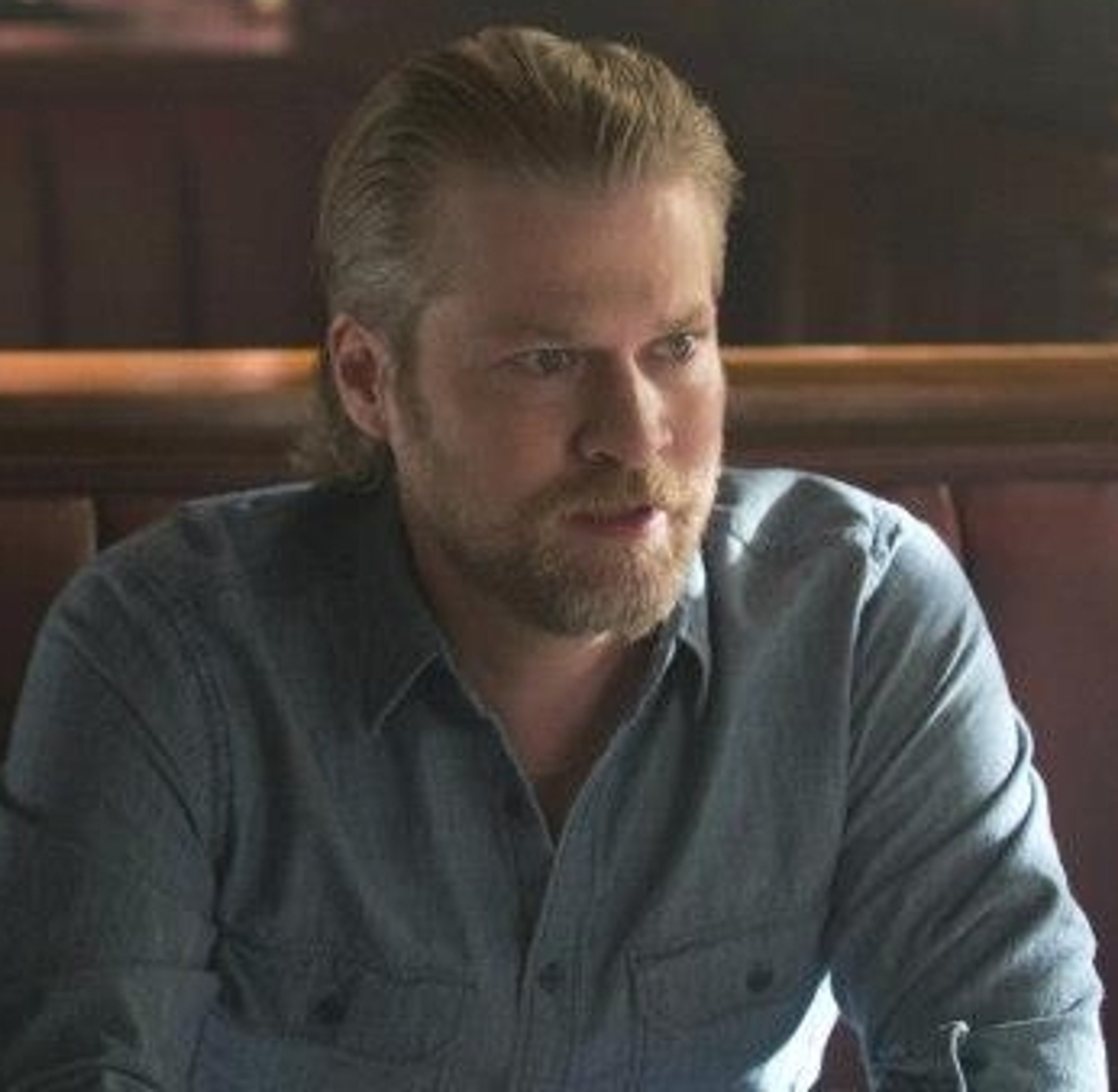 A middle-aged man with short blonde hair and a beard, wearing a gray button-up shirt and looking serious.