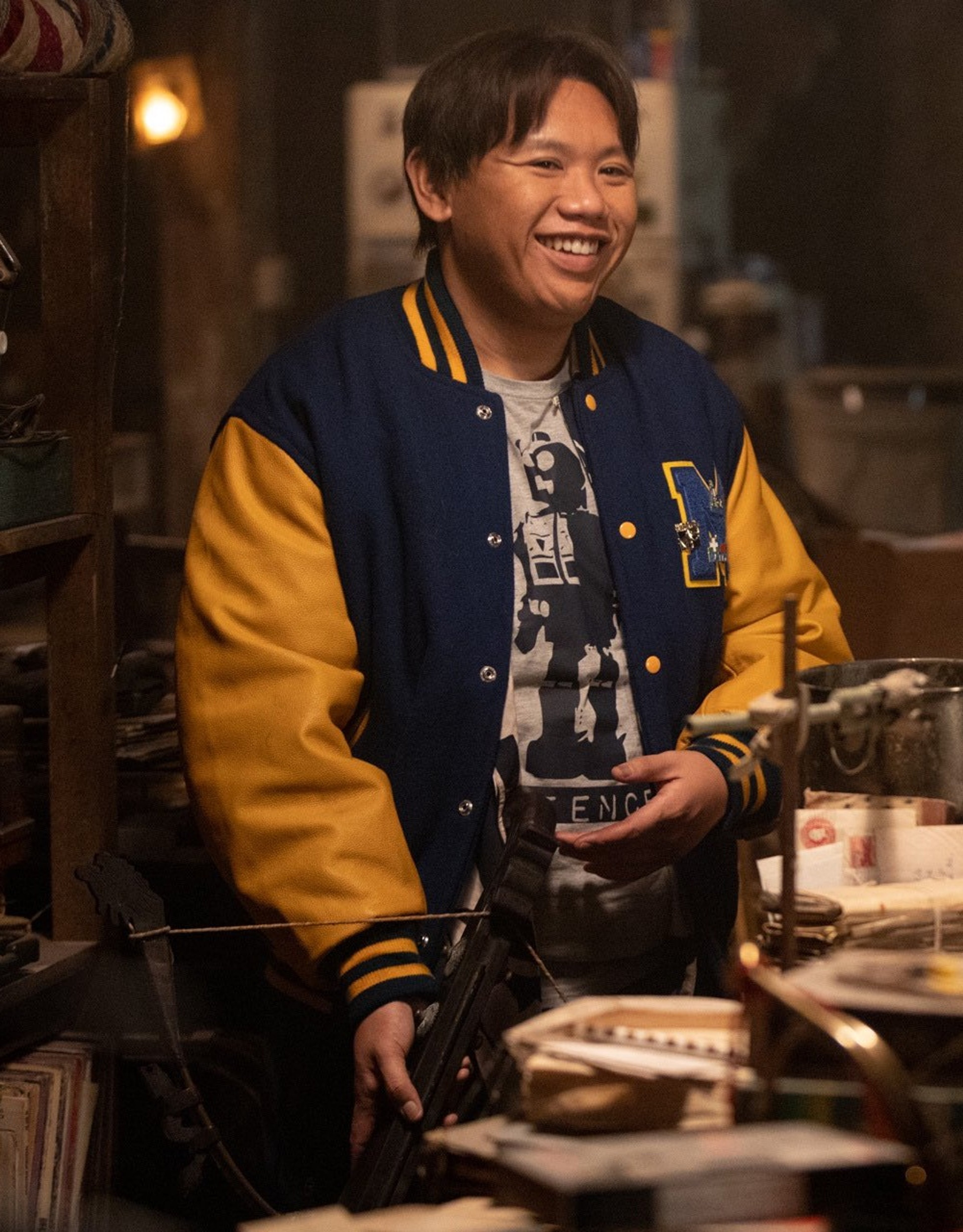 A young Asian man with a friendly, enthusiastic expression wearing a navy blue and yellow varsity jacket