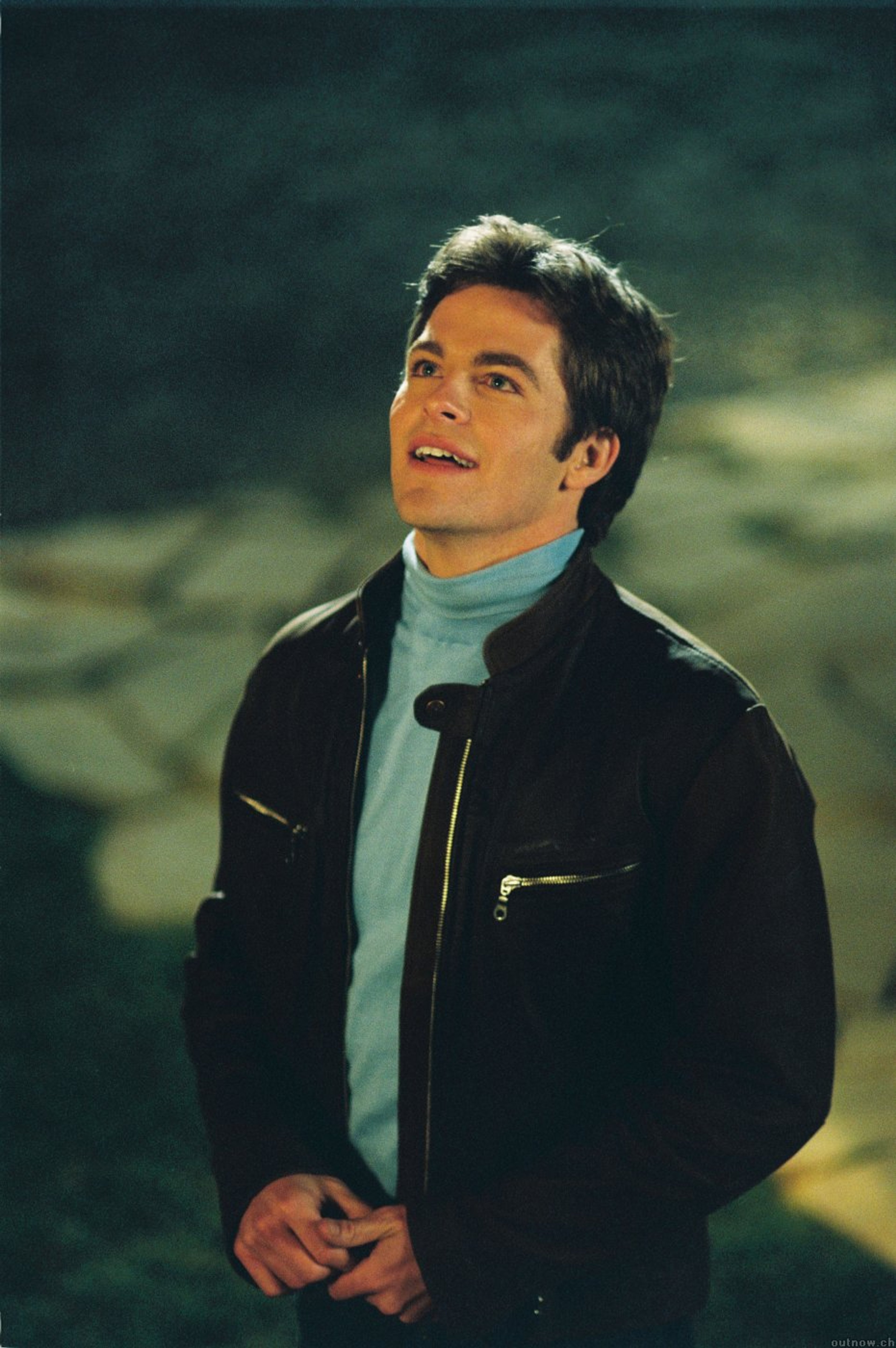 A man with dark hair and a confident expression, wearing a black jacket and light blue scarf.