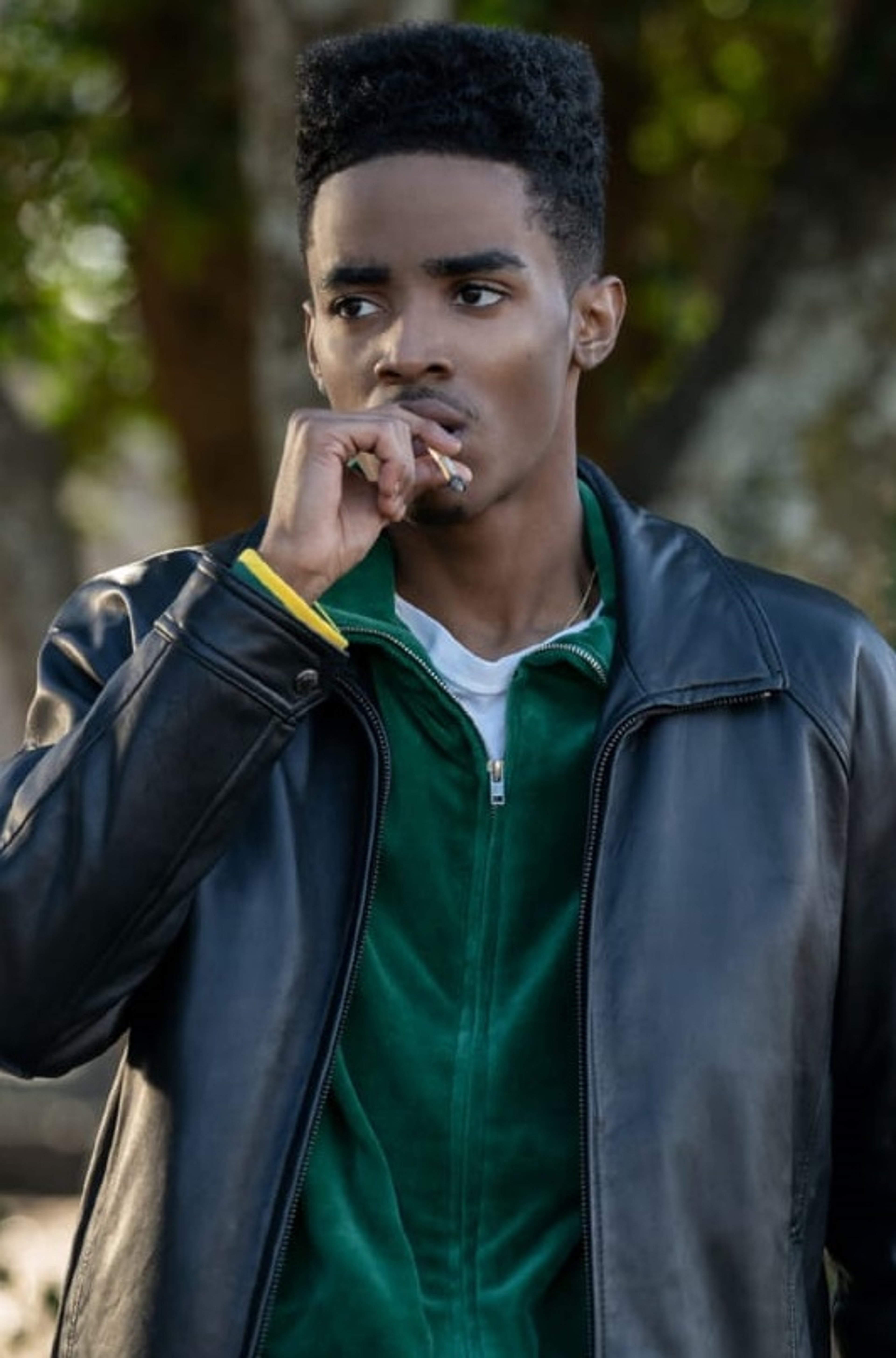 A young Black man with short, curly hair wearing a green jacket and holding his hand to his mouth in a thoughtful gesture.