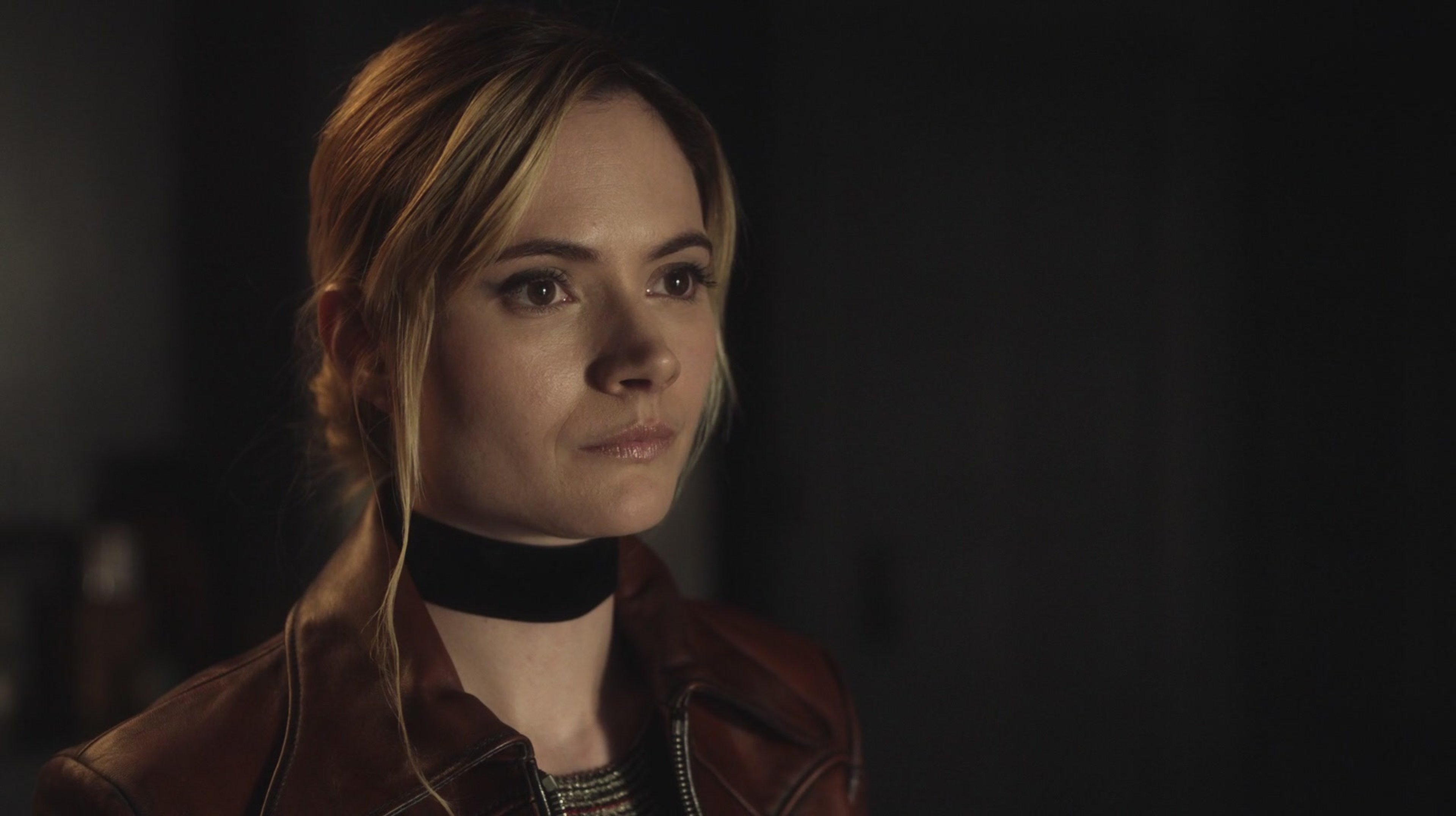 A woman with short brown hair wearing a red choker and a dark outfit, with an intense expression on her face.