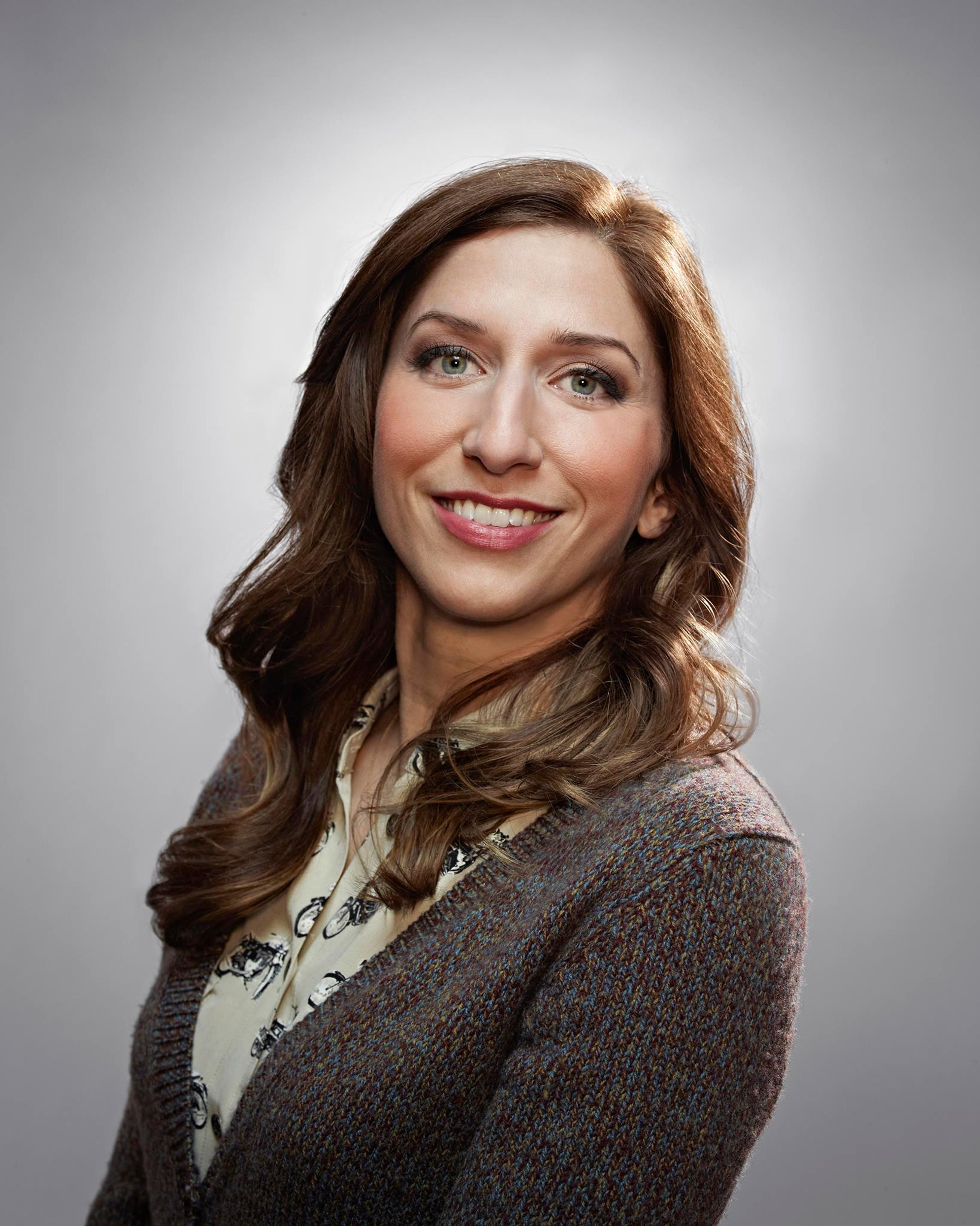 A smiling woman with long brown hair wearing a gray sweater