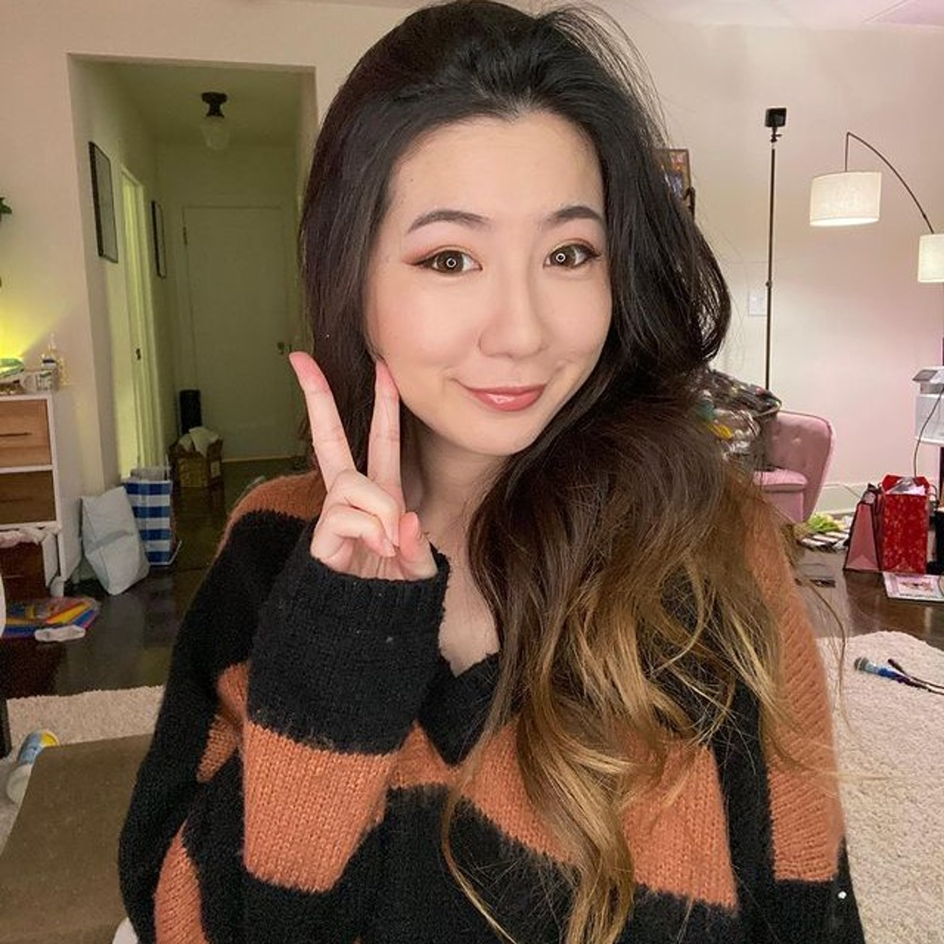 A young woman with long dark hair making a peace sign gesture while wearing a black and orange striped sweater