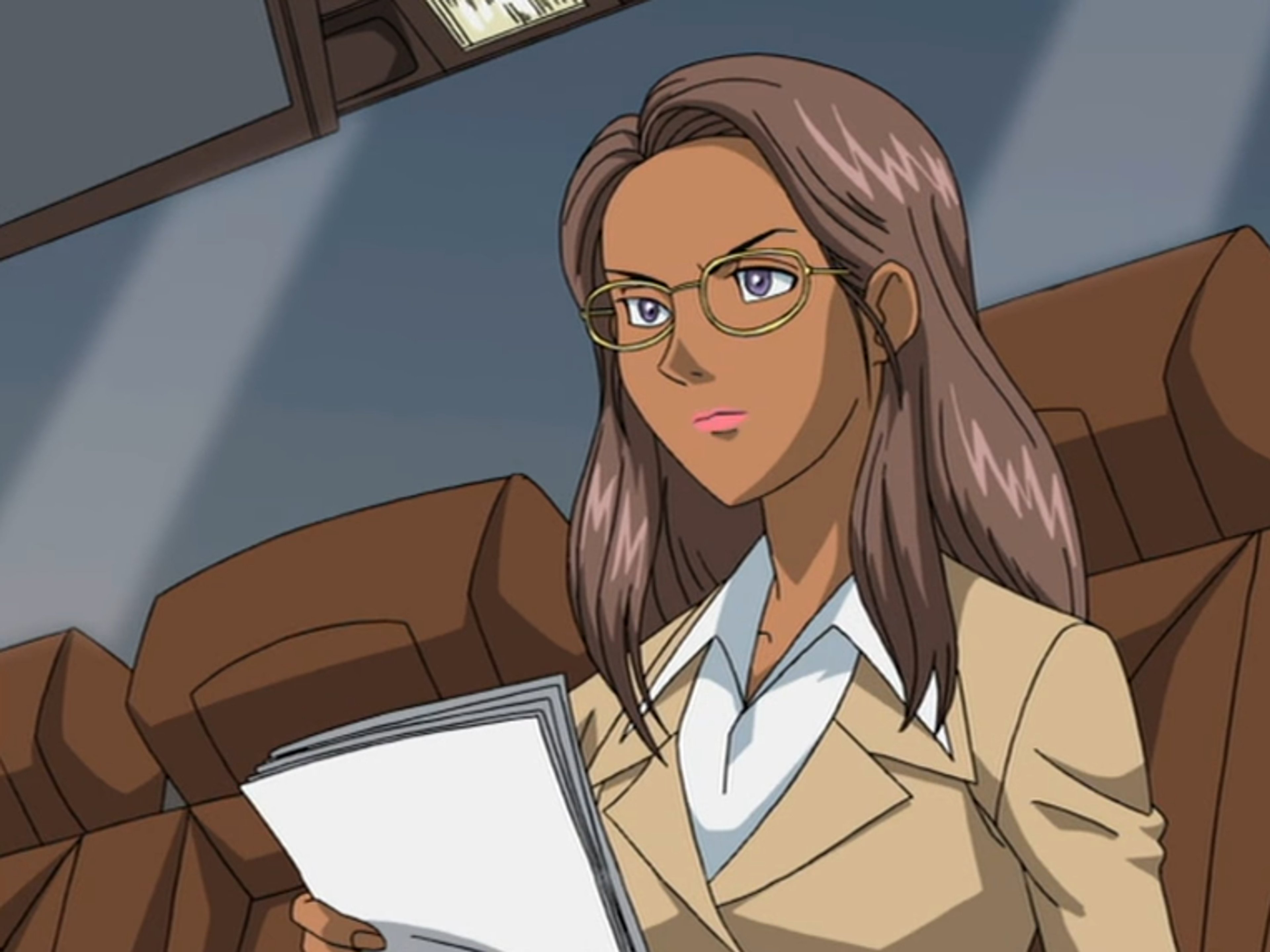 A professional-looking woman in a tan suit and glasses, sitting at a desk in an office setting.