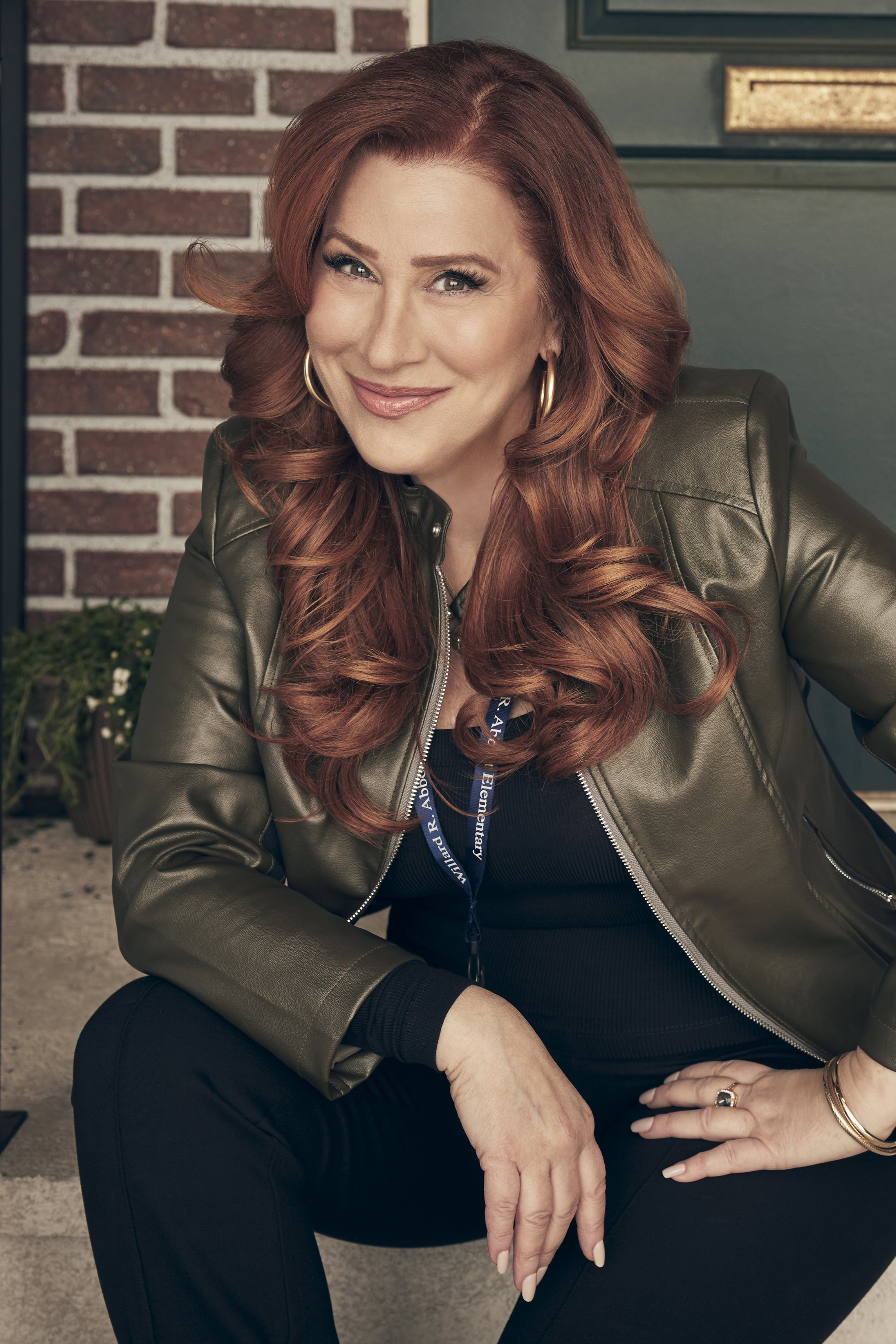 A woman with long, wavy red hair wearing a black leather jacket, sitting in front of a brick wall and smiling.