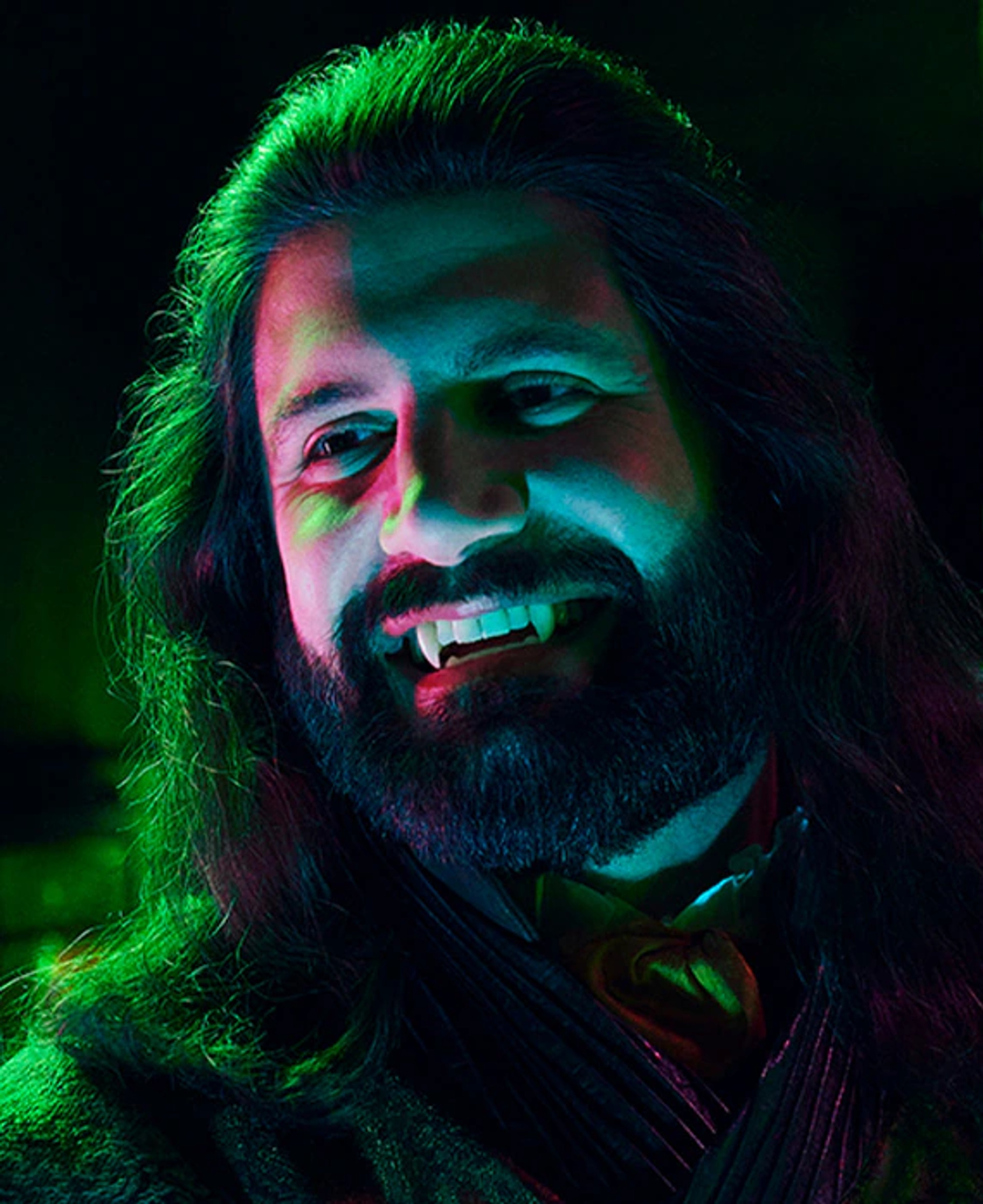 A man with long dark hair and a beard wearing a dark robe or cloak, with dramatic green and blue lighting