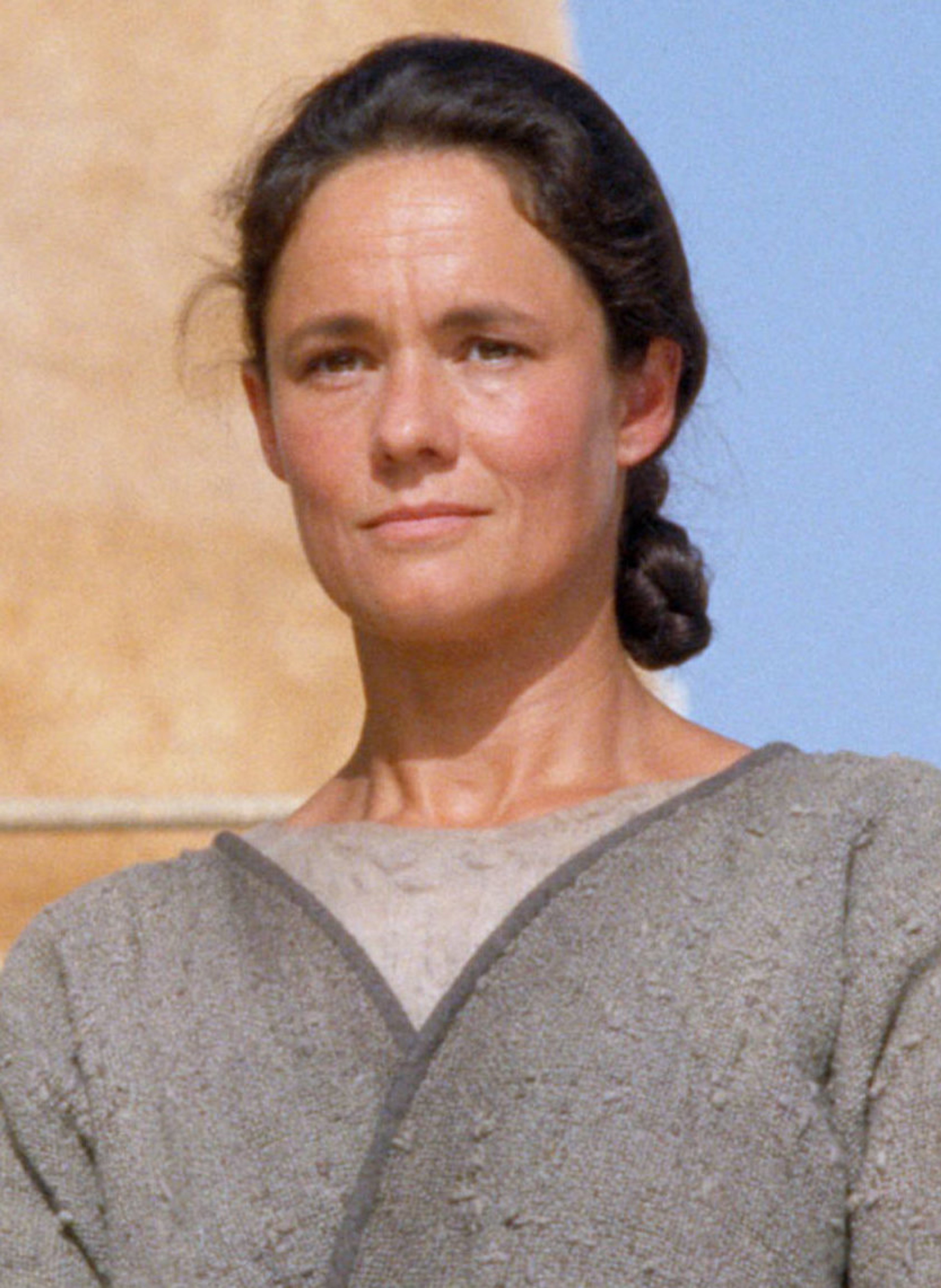 A middle-aged woman with dark hair in a braid, wearing a gray tunic, in a desert-like setting.