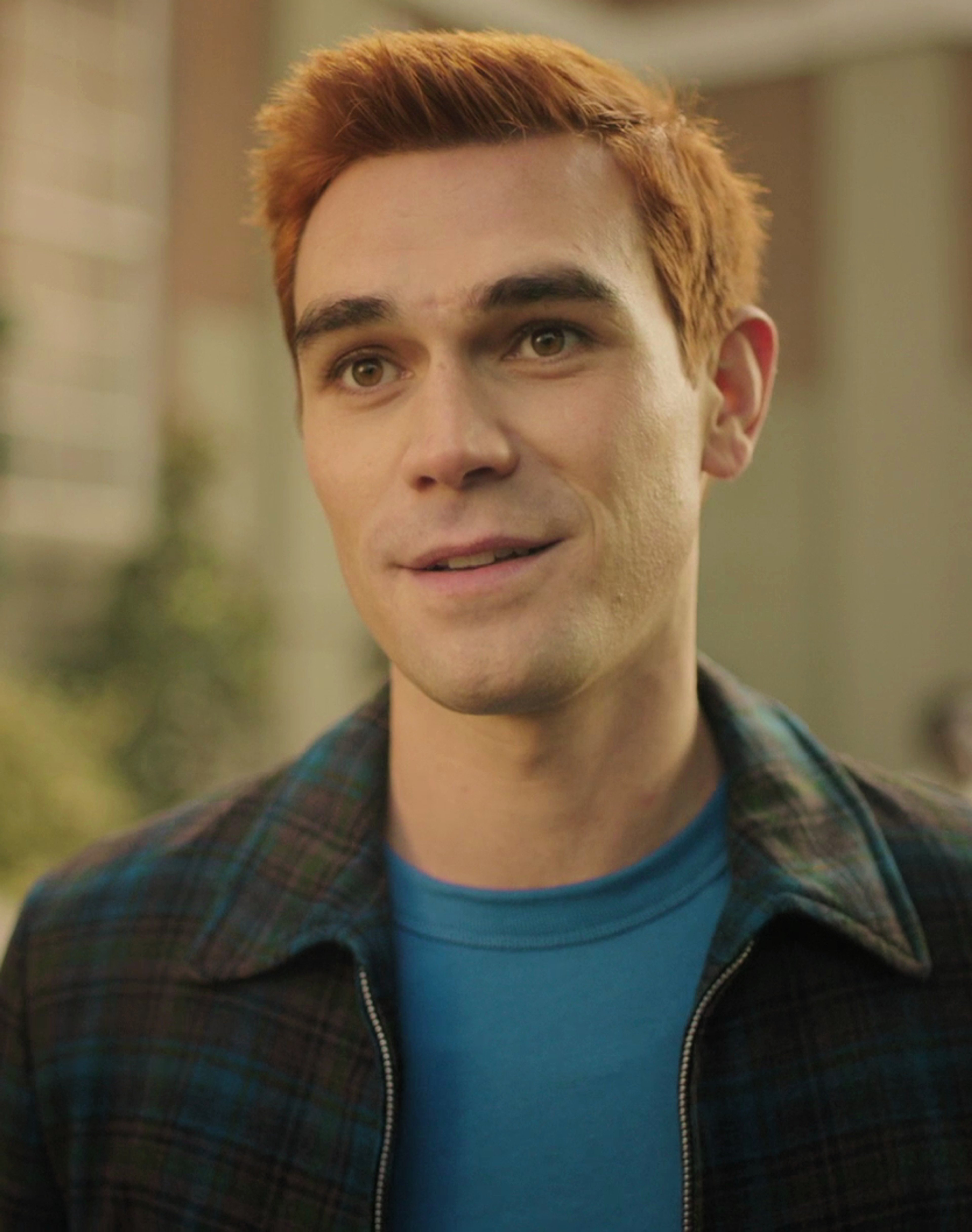 A young man with reddish-brown hair and blue eyes wearing a plaid jacket and blue t-shirt