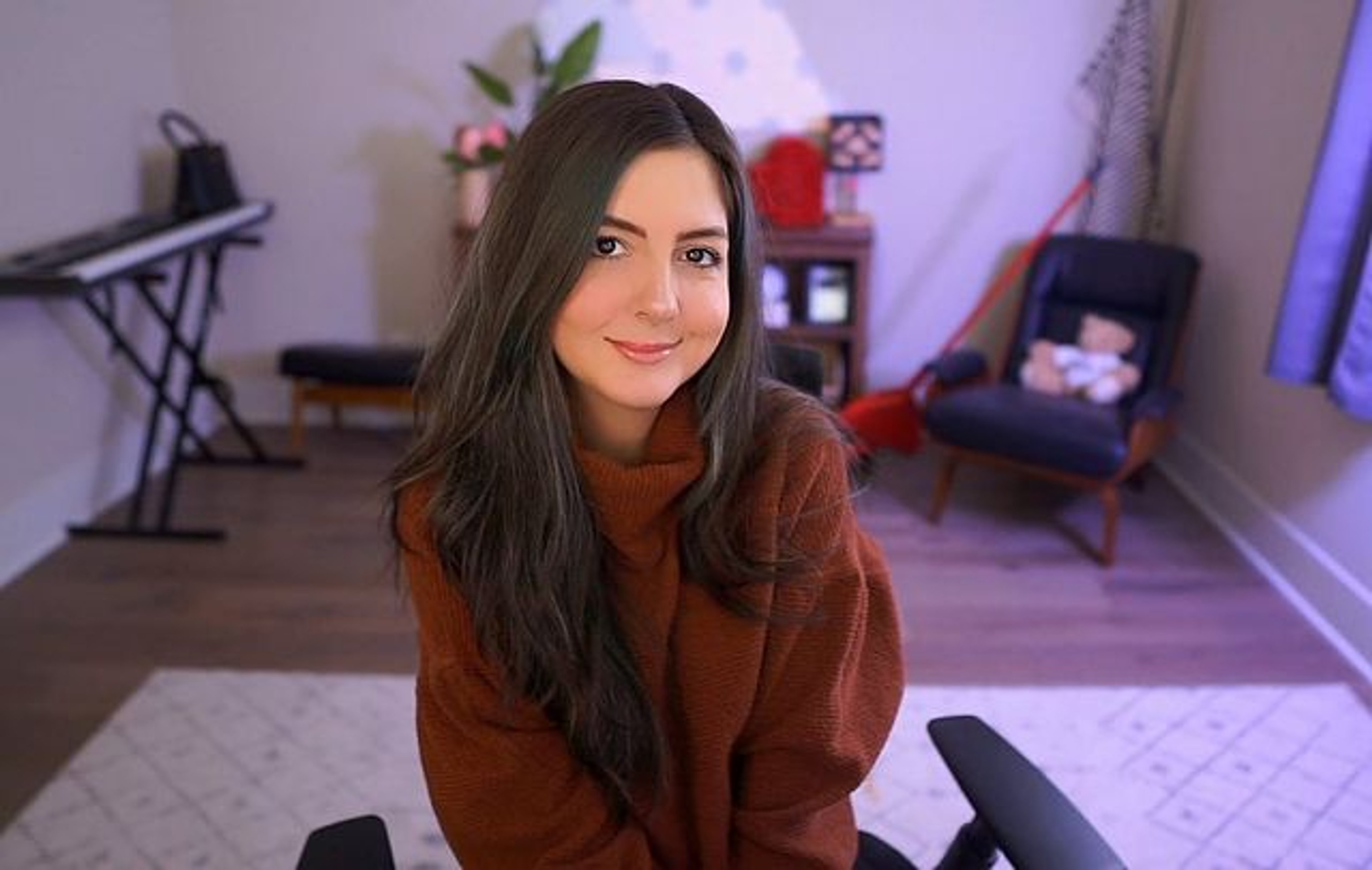 A young woman with long dark hair sitting in a room, wearing a brown sweater and smiling.
