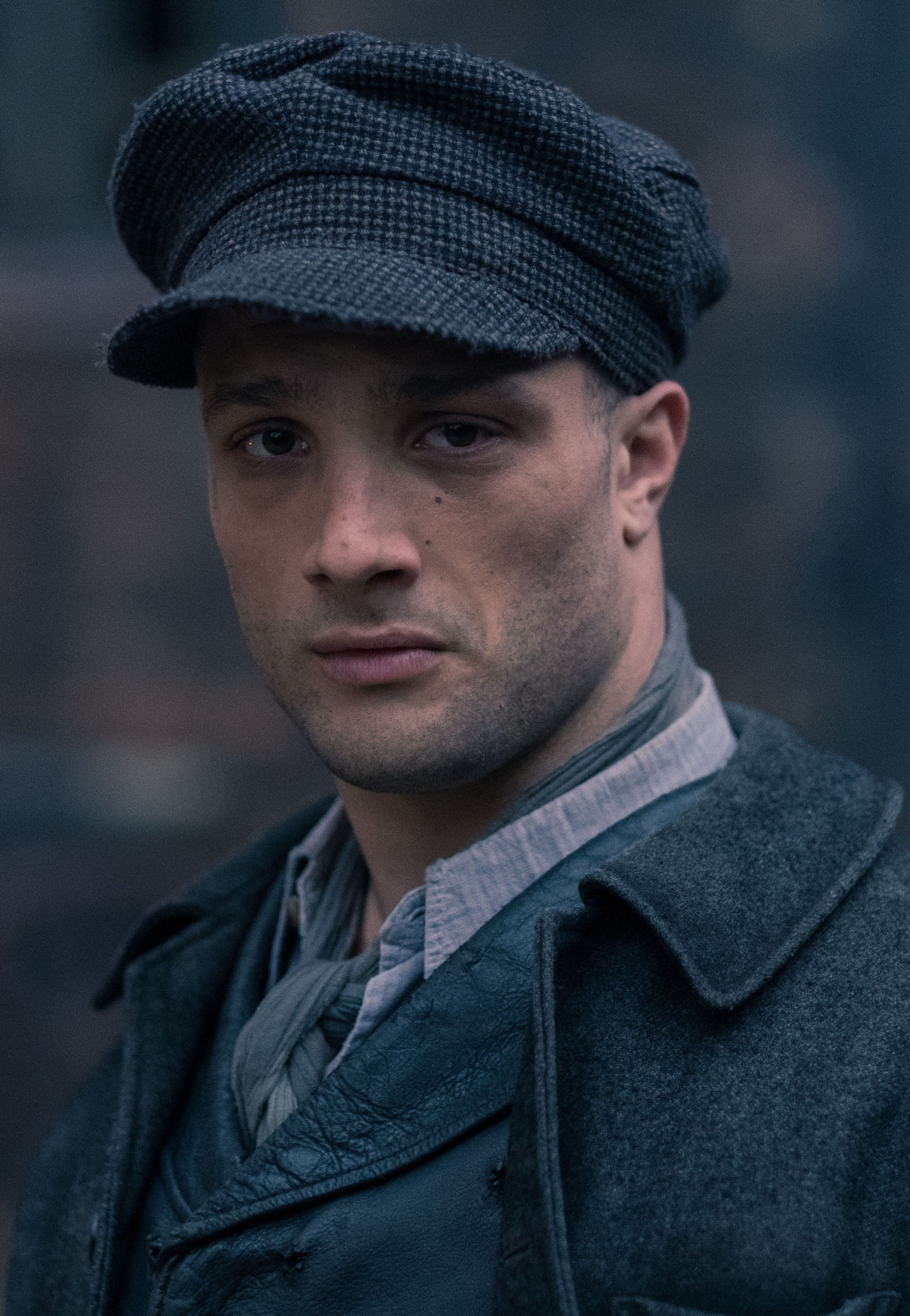 A man with a serious expression wearing a dark coat and flat cap