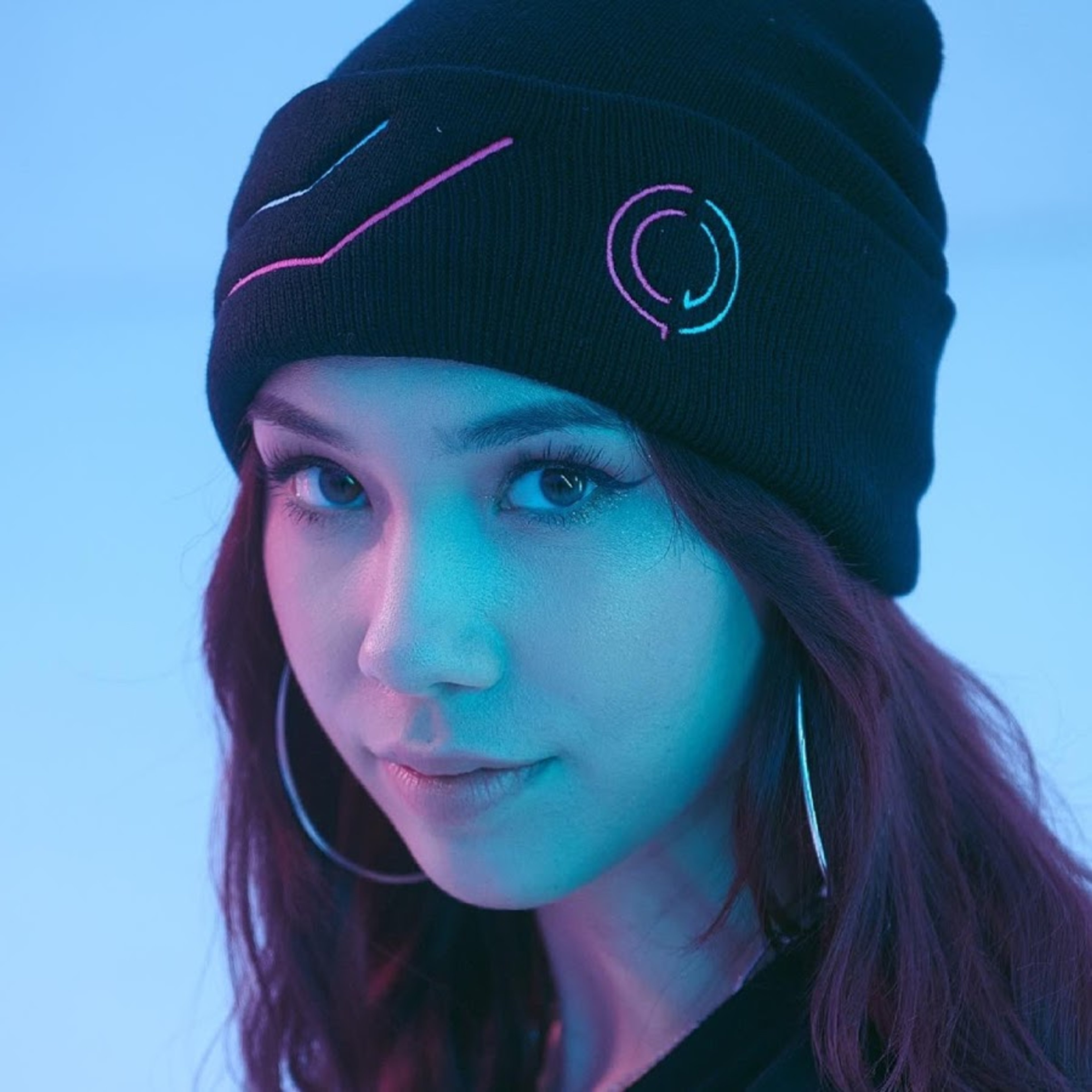 A young woman with long dark hair wearing a black beanie with a colorful design, looking directly at the camera with a friendly expression.
