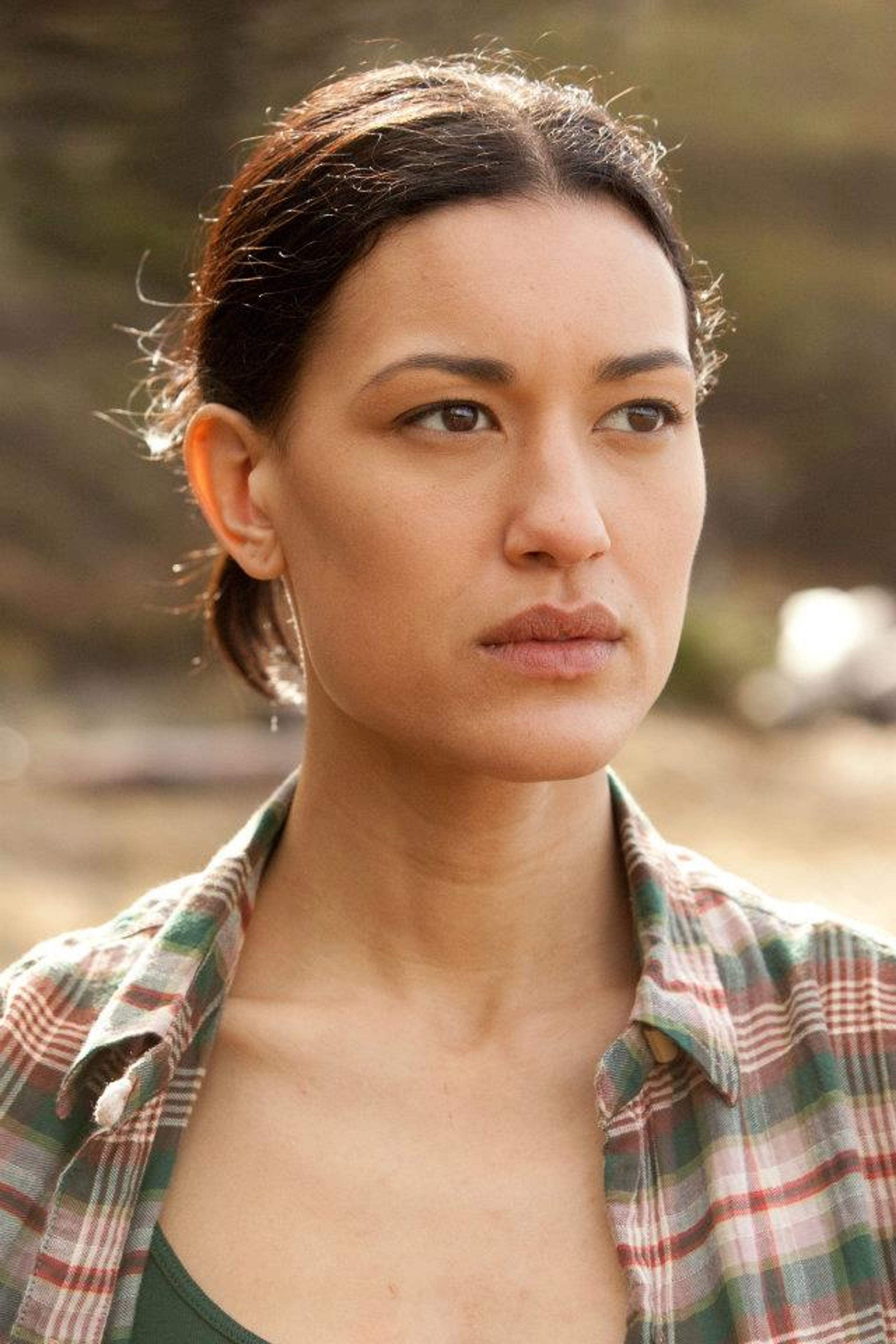 A young woman with dark hair wearing a plaid shirt in an outdoor setting.