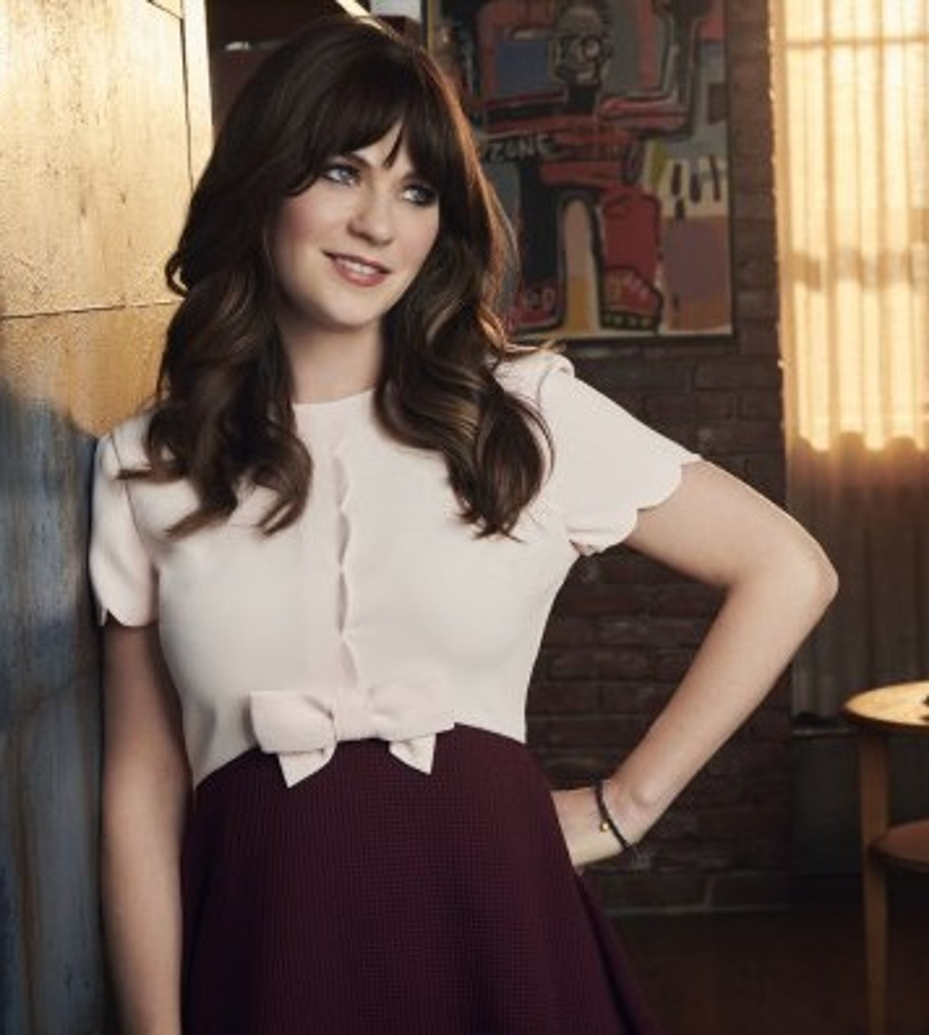 A young woman with dark curly hair wearing a white blouse and burgundy skirt, standing in a loft or apartment setting.