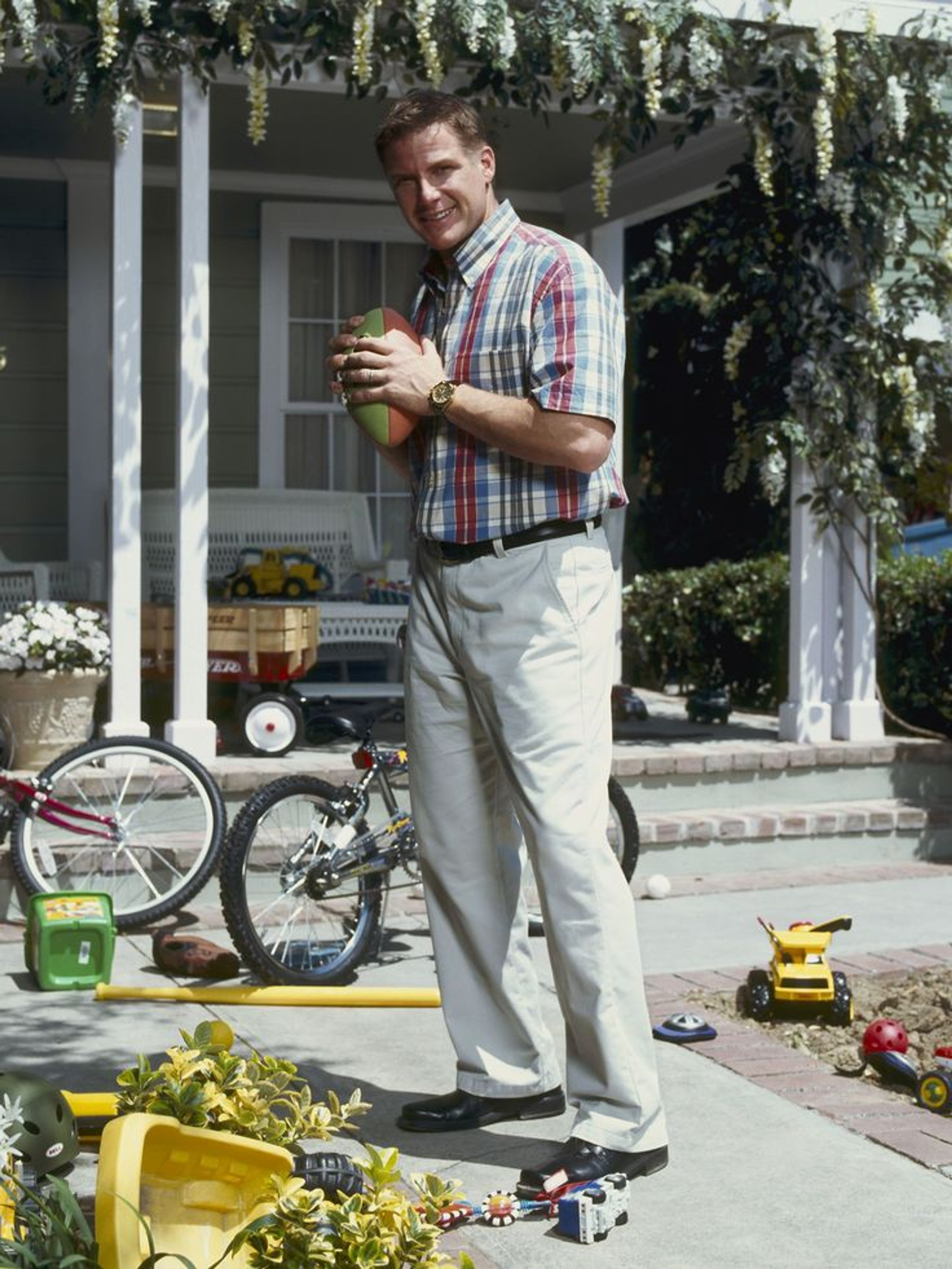 A middle-aged man standing in a garden or yard, holding a drink and smiling.