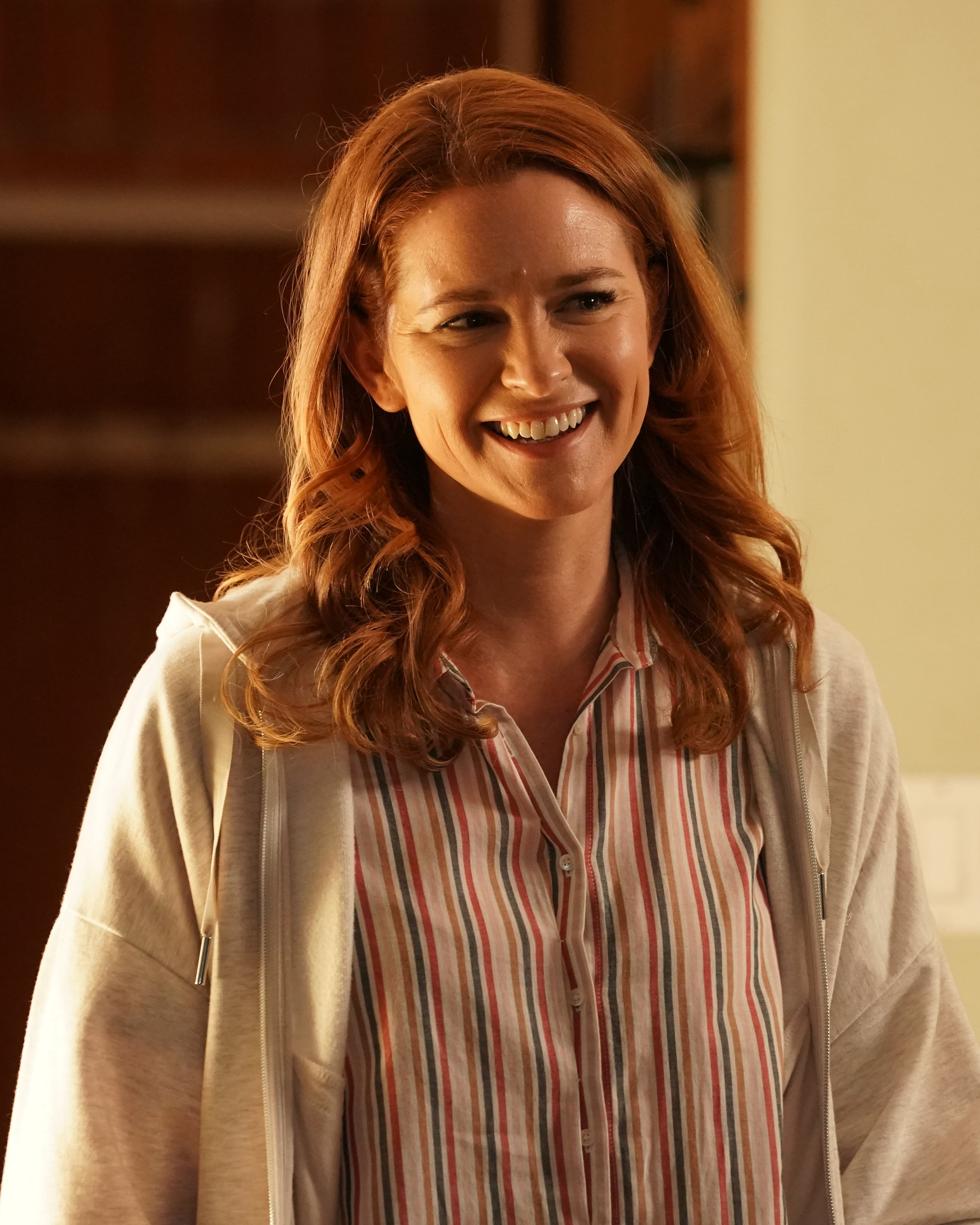 A smiling woman with long, wavy red hair wearing a white striped shirt