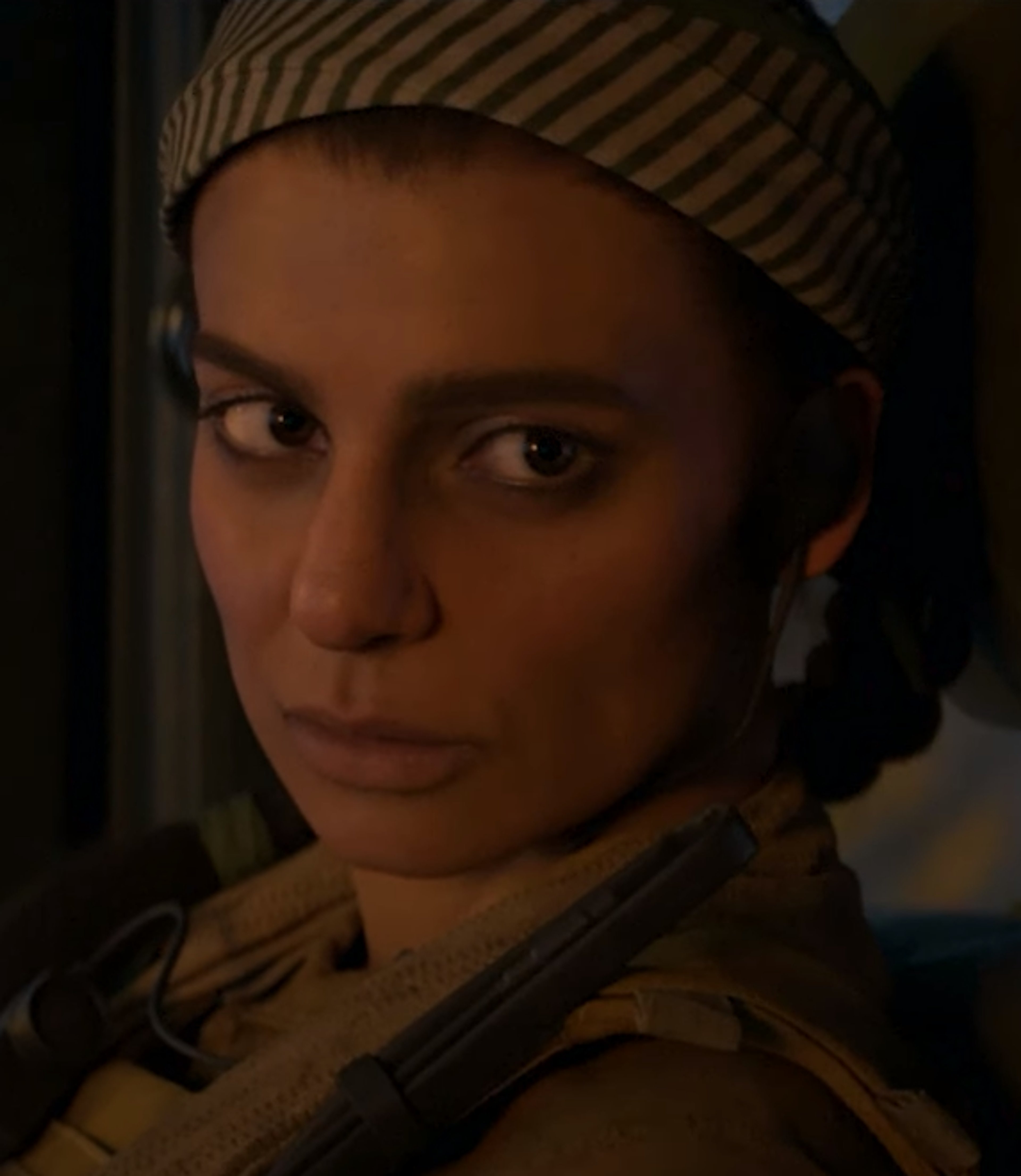 A woman with dark hair wearing a tan military-style hat and jacket, with a serious expression on her face.