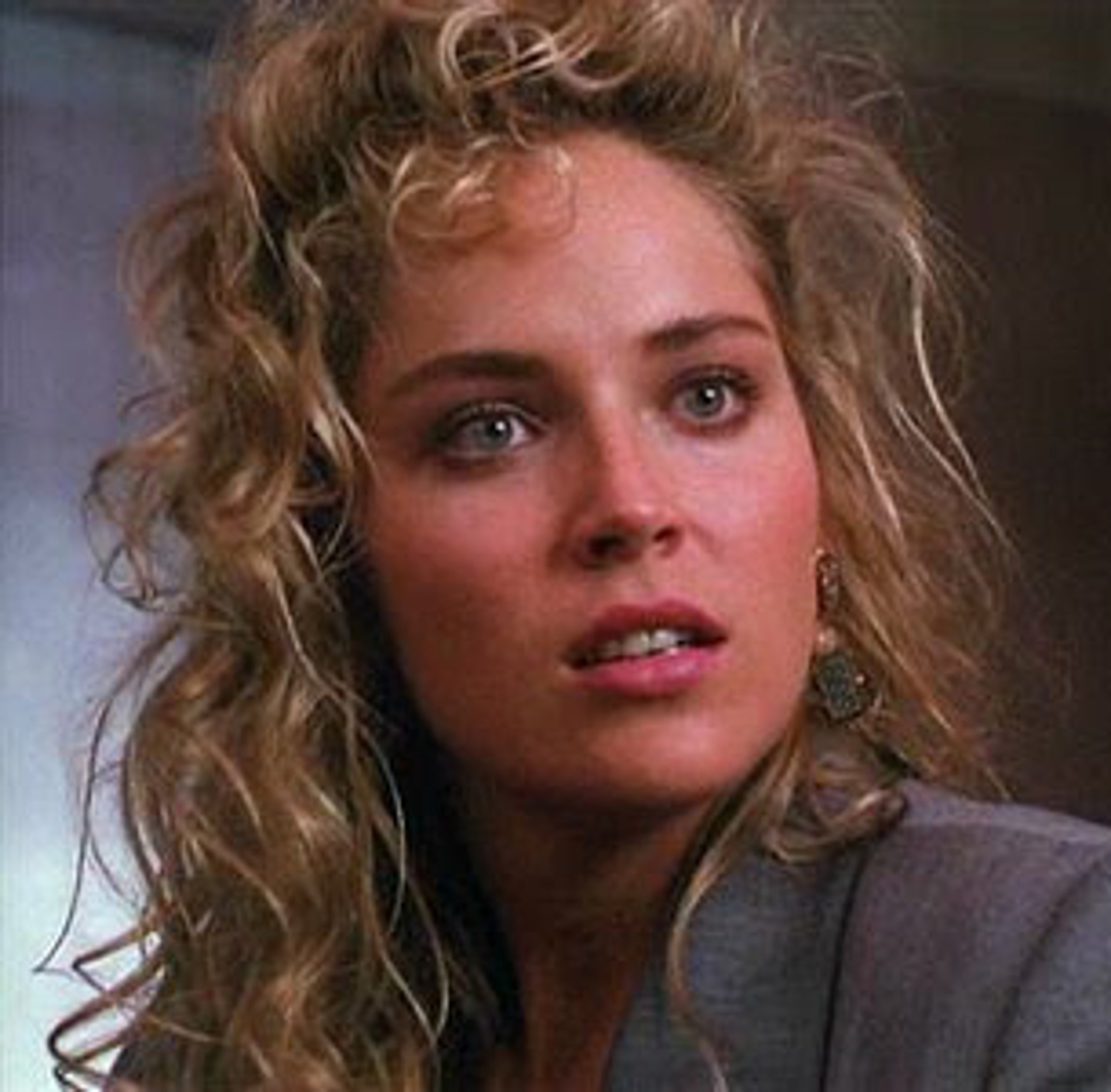 A woman with curly blonde hair and a serious expression, wearing a gray jacket or shirt.