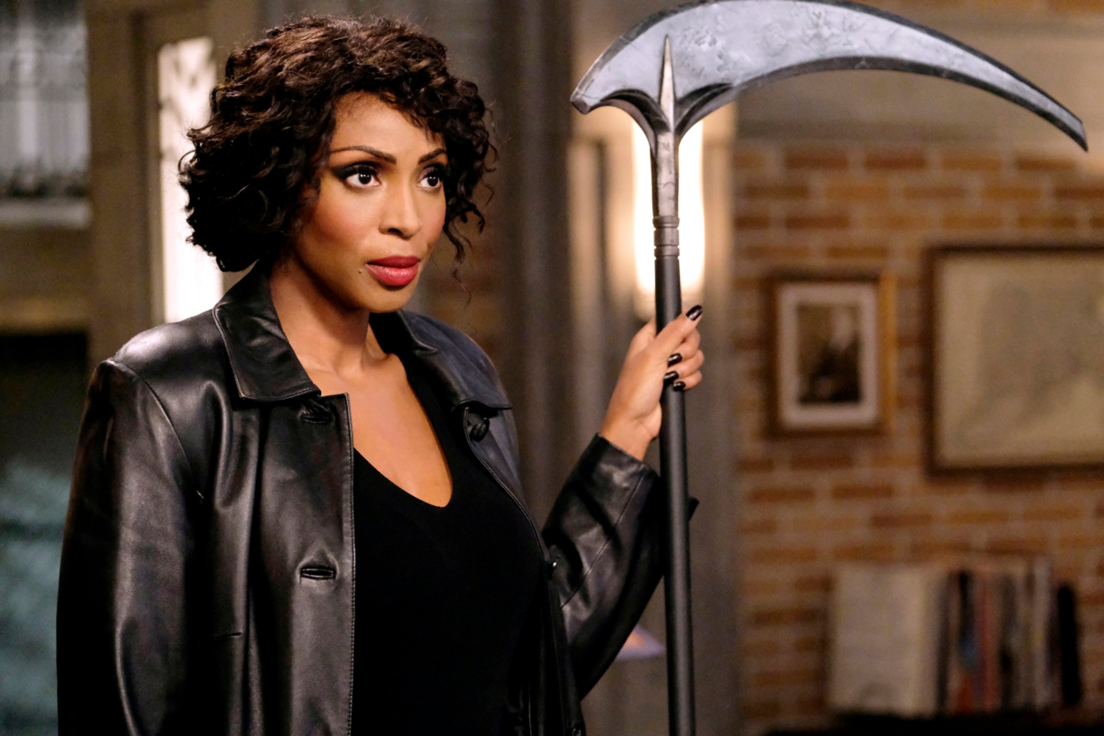 A woman with curly dark hair wearing a black leather jacket and holding a large scythe-like weapon