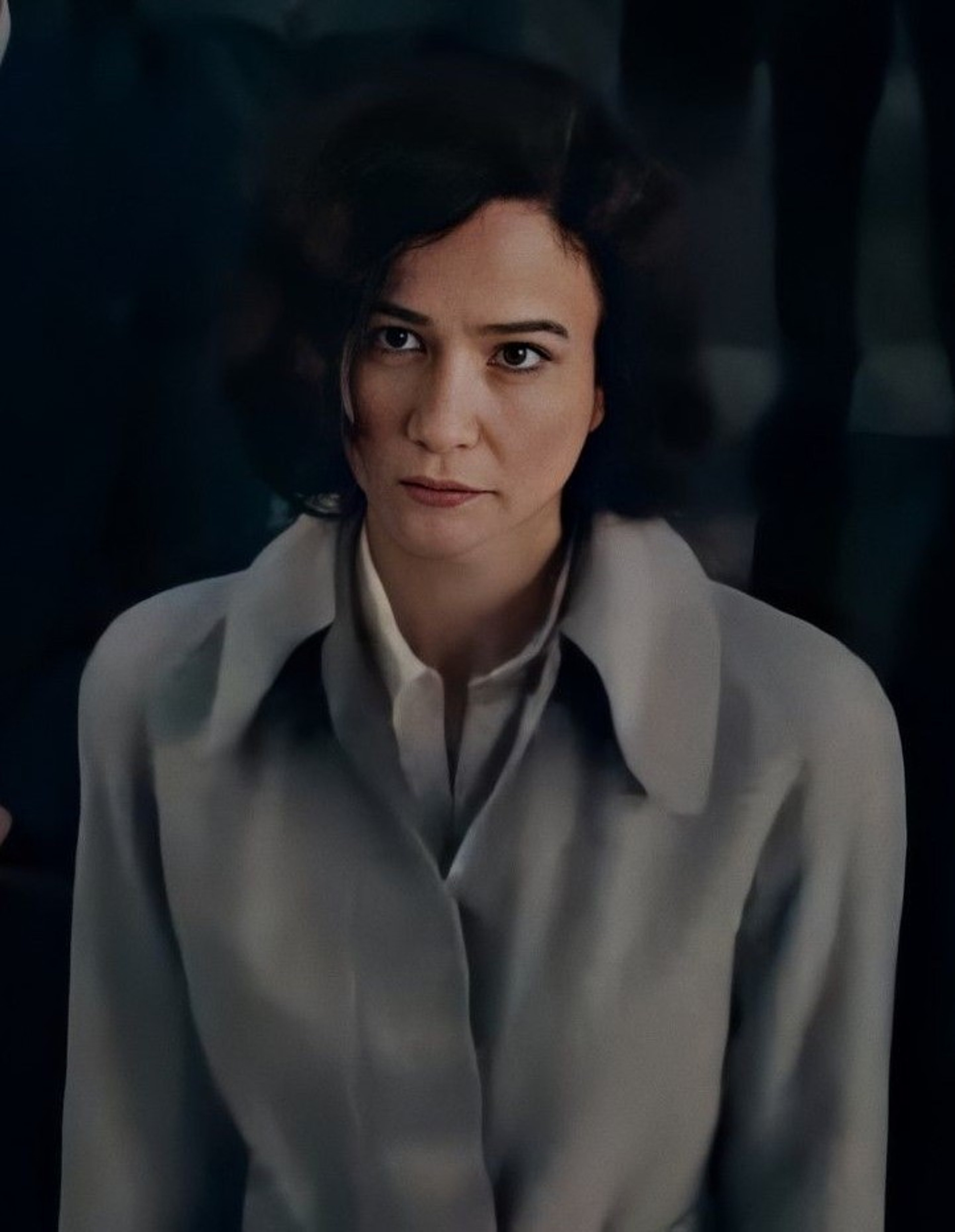 A woman with dark hair wearing a light-colored coat, looking directly at the camera with a serious expression.