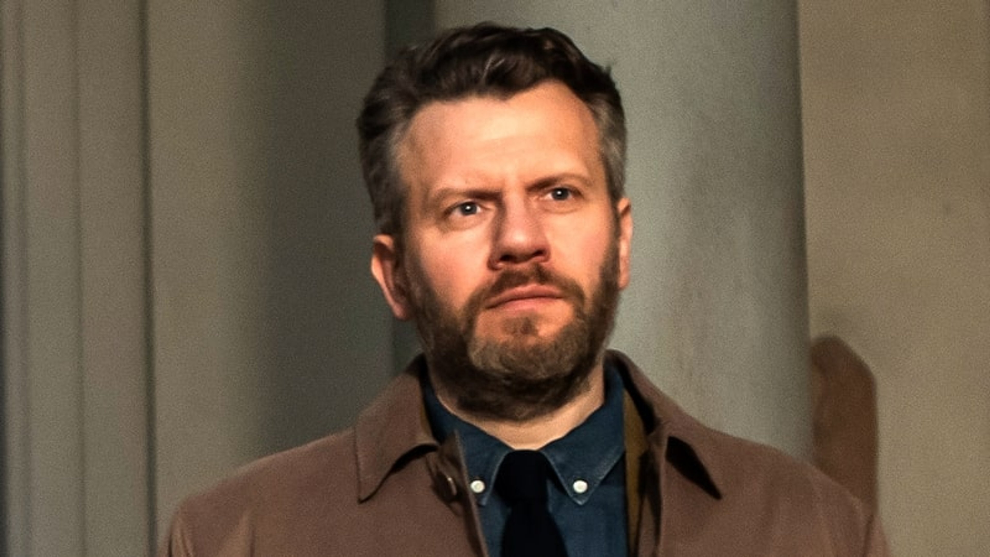 A middle-aged man with a beard and short dark hair, wearing a brown jacket and blue shirt, looking directly at the camera.