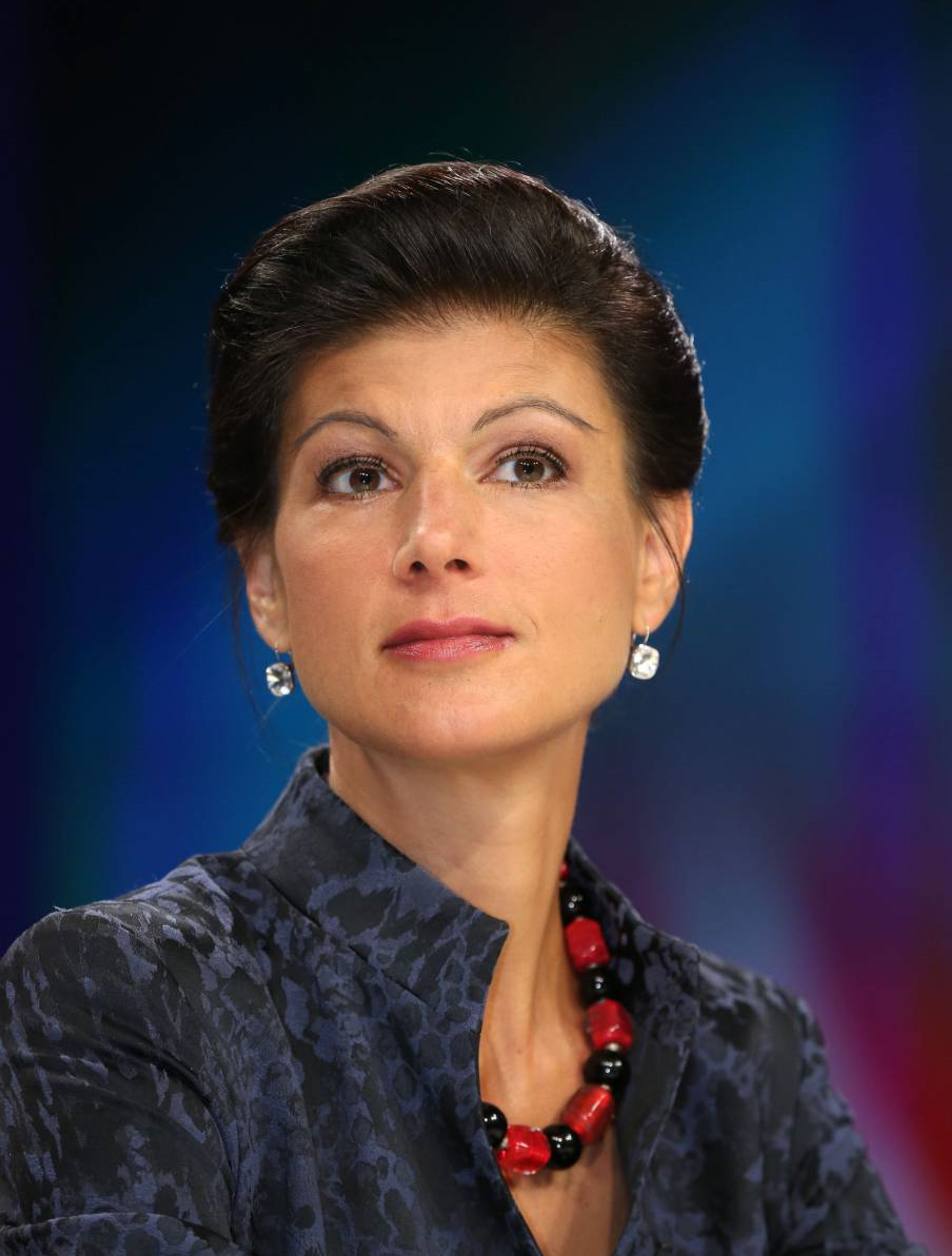 Portrait of a serious-looking middle-aged woman with dark hair and a red bead necklace against a blue background.