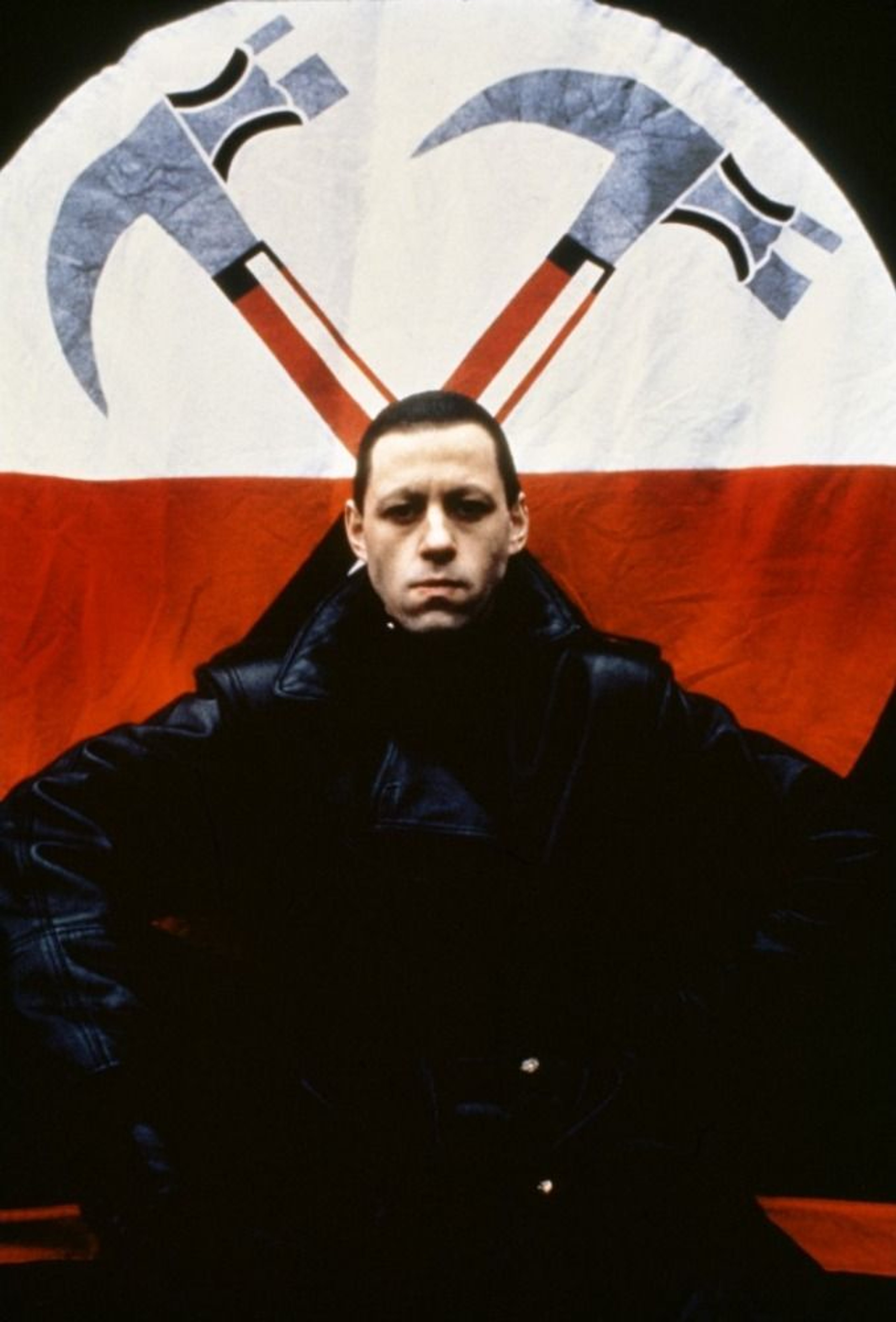 A man with a serious expression in front of large hammers or axes