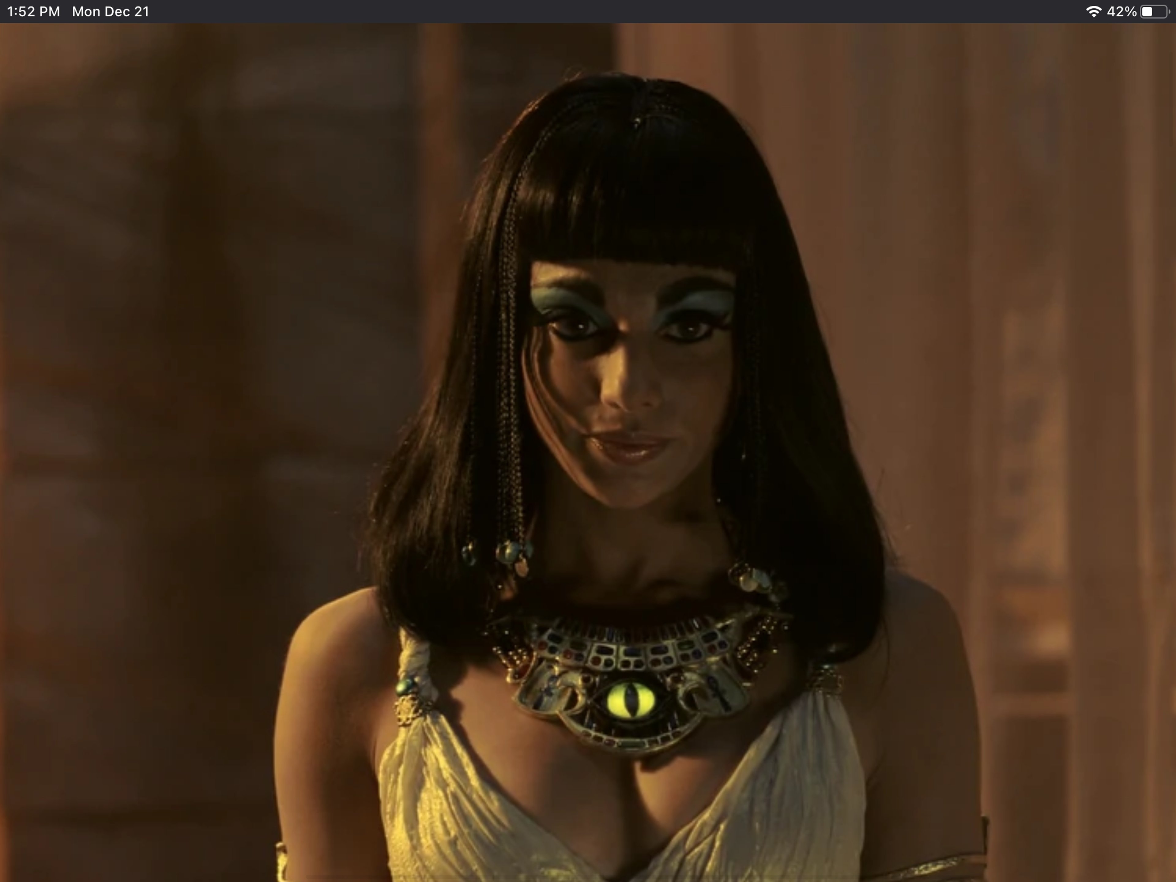 A woman with long dark hair and striking blue eyes wearing a gold necklace with a cat's eye pendant, conveying a sense of ancient Egyptian royalty and mysticism.