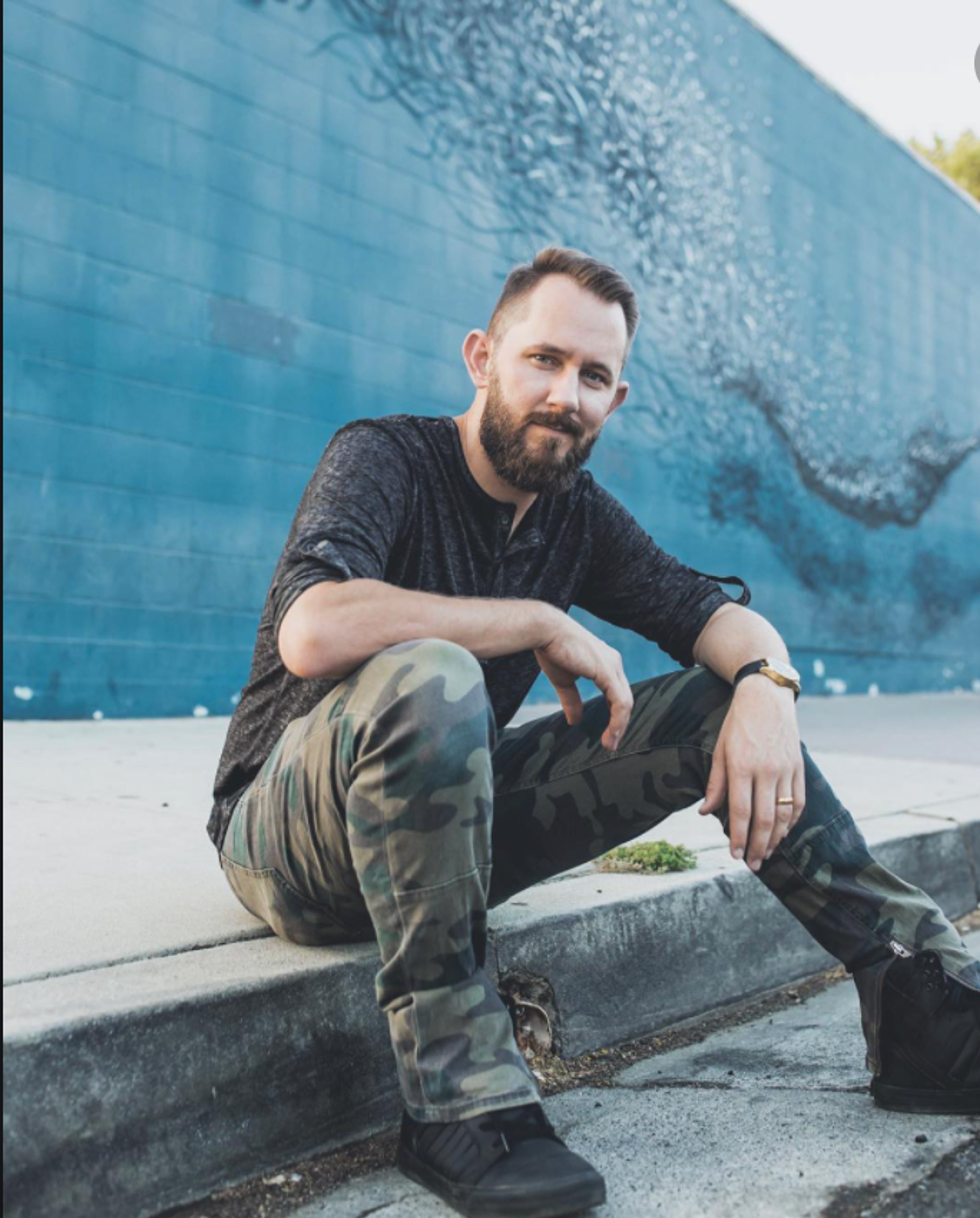 A man with a beard sitting on a concrete ledge in an urban setting