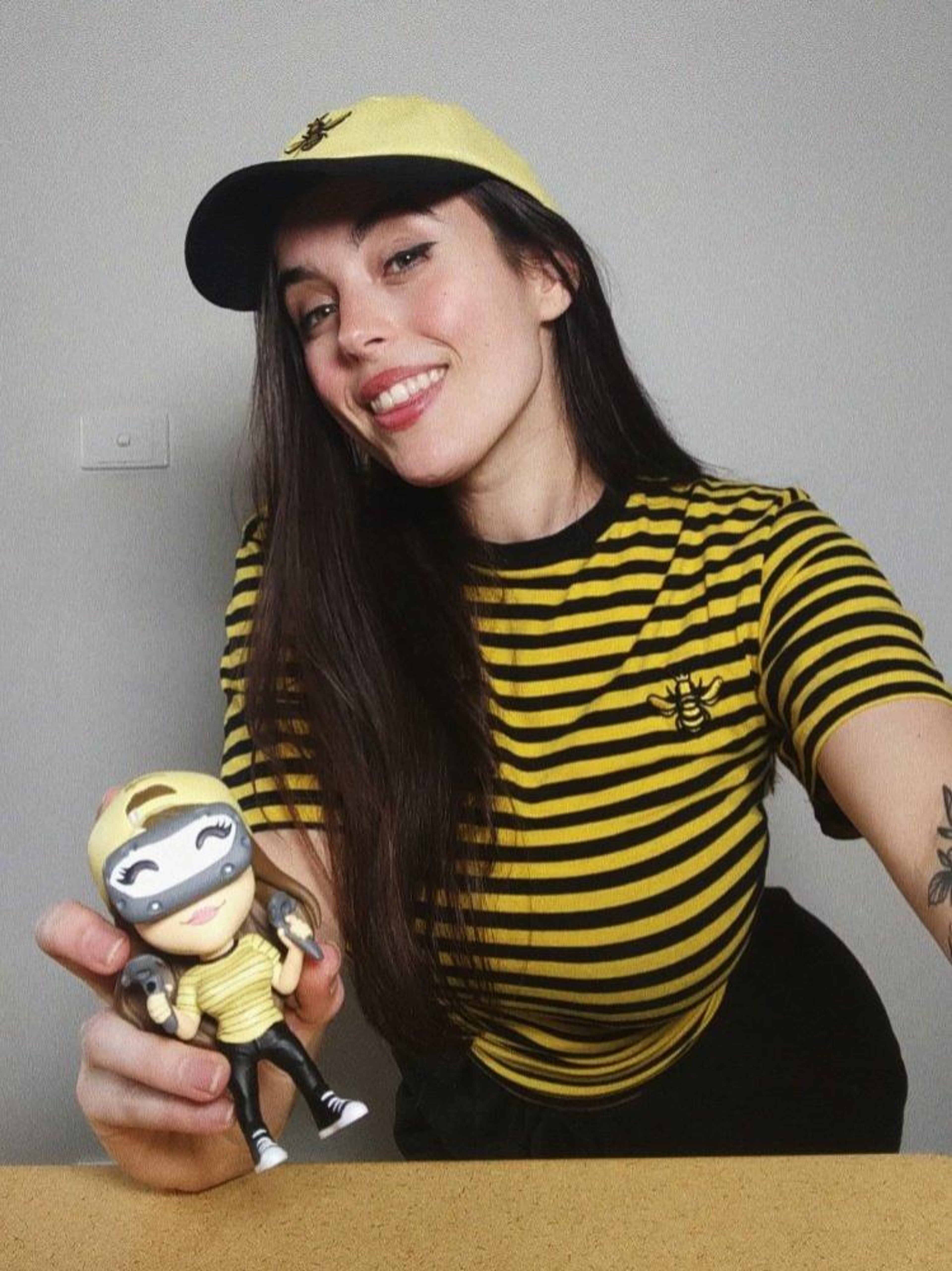 A young woman with long dark hair wearing a yellow and black striped shirt and hat, smiling and holding a plush toy of a cartoon monkey or ape.