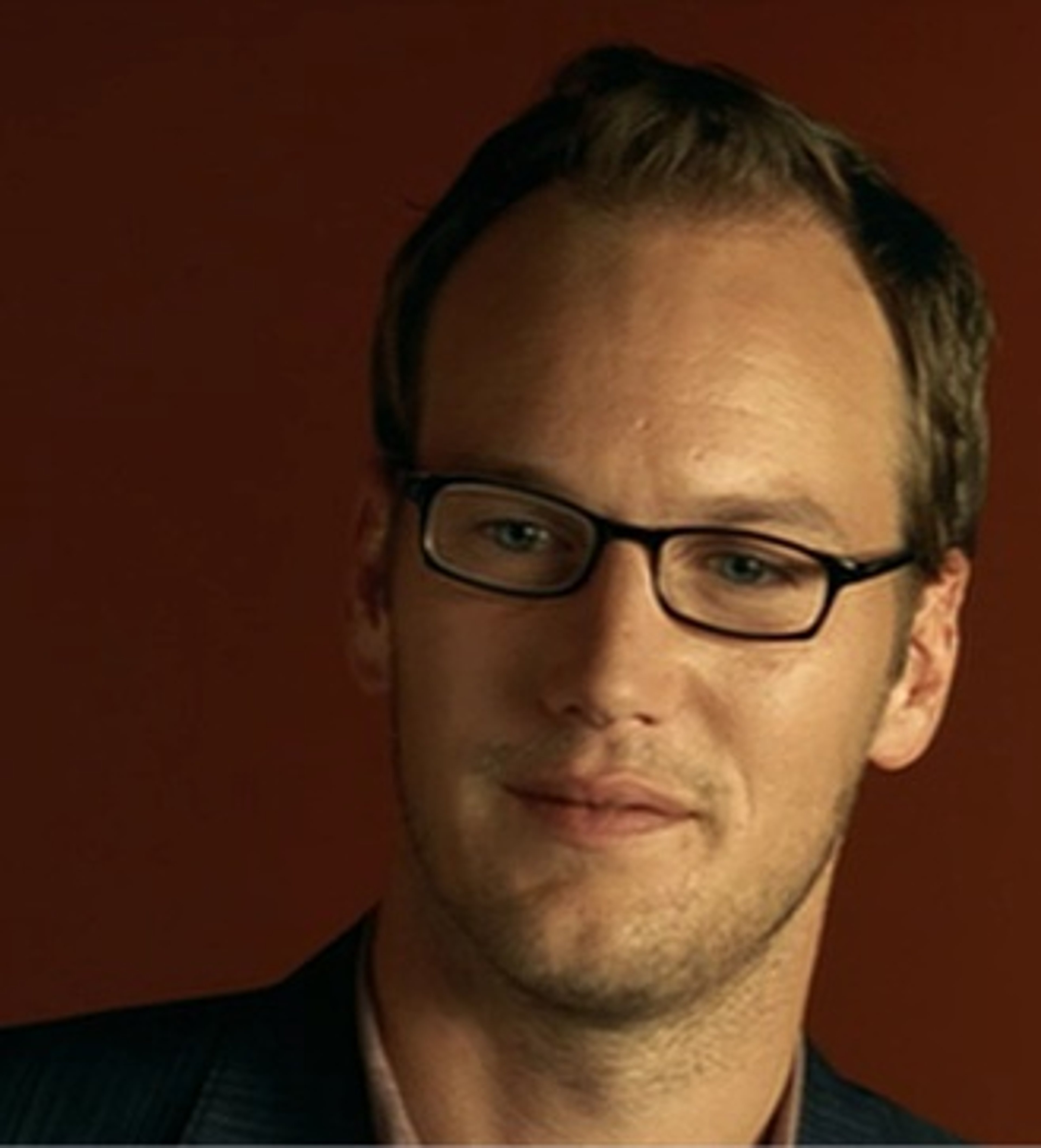 A man in his 30s with short brown hair and glasses, wearing a black suit jacket and smiling.