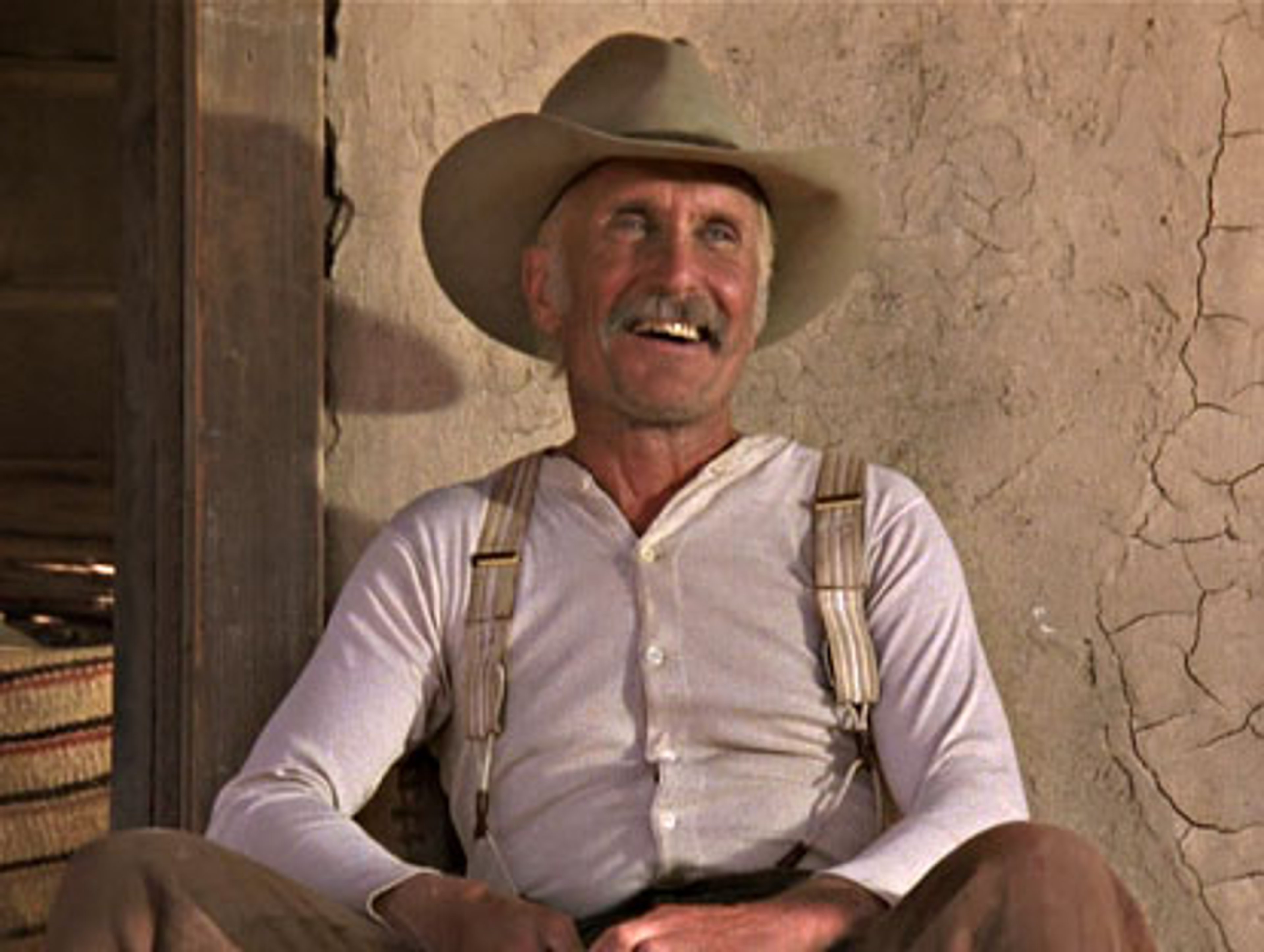 An older, rugged-looking cowboy sitting in a wooden building