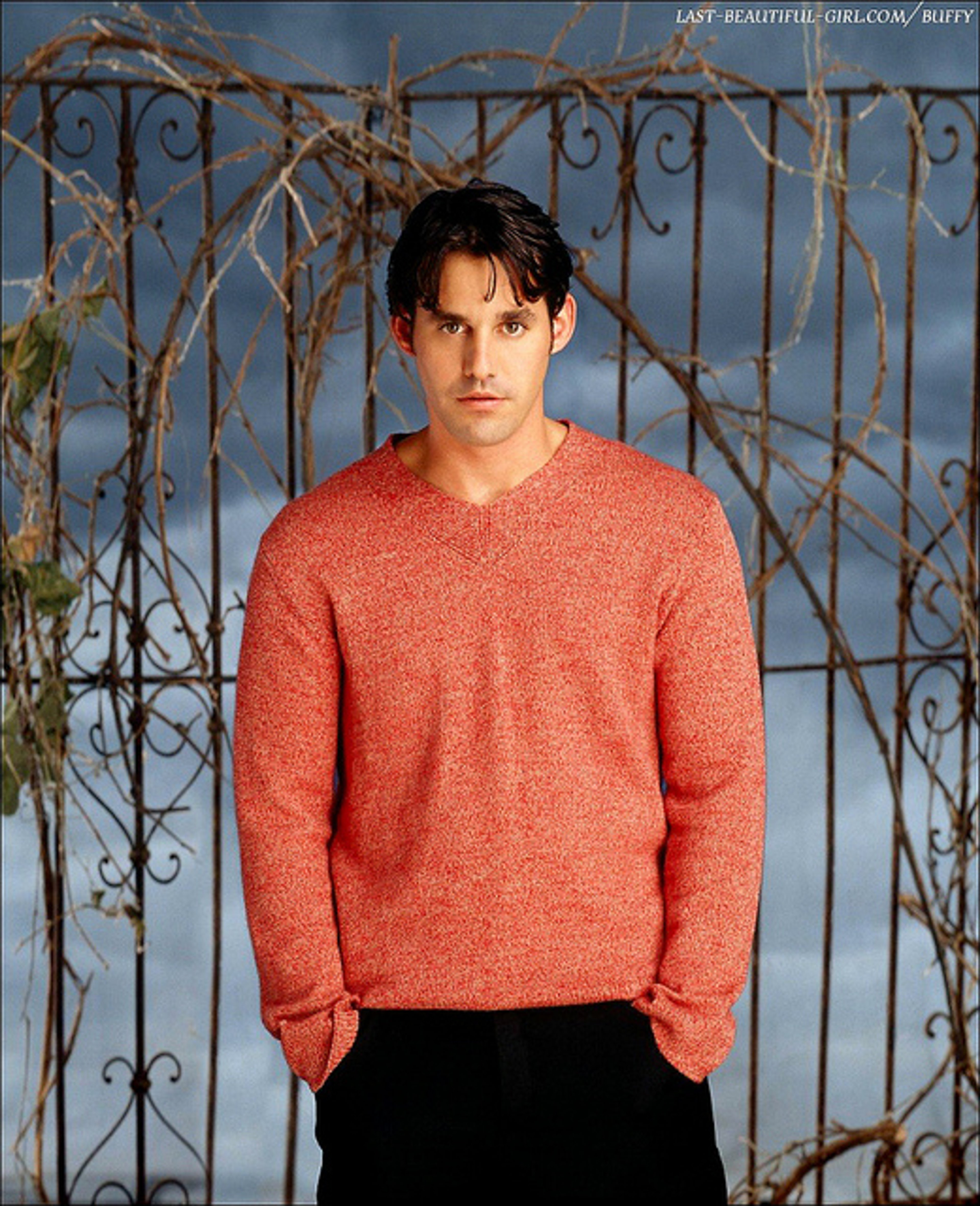 A young man with dark hair wearing a red sweater standing in front of a wrought iron gate or fence.