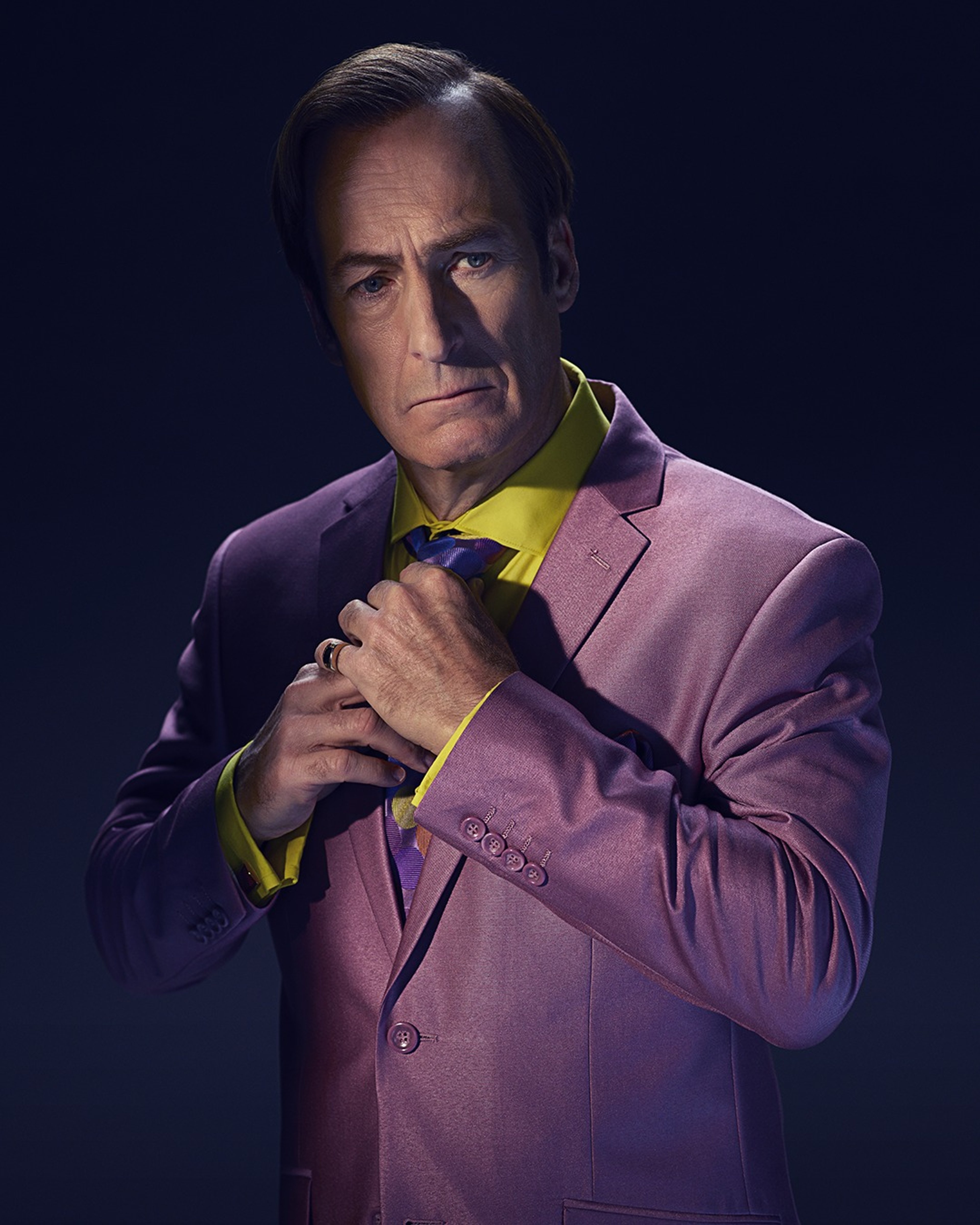 A man in a purple suit jacket and yellow shirt with a serious expression