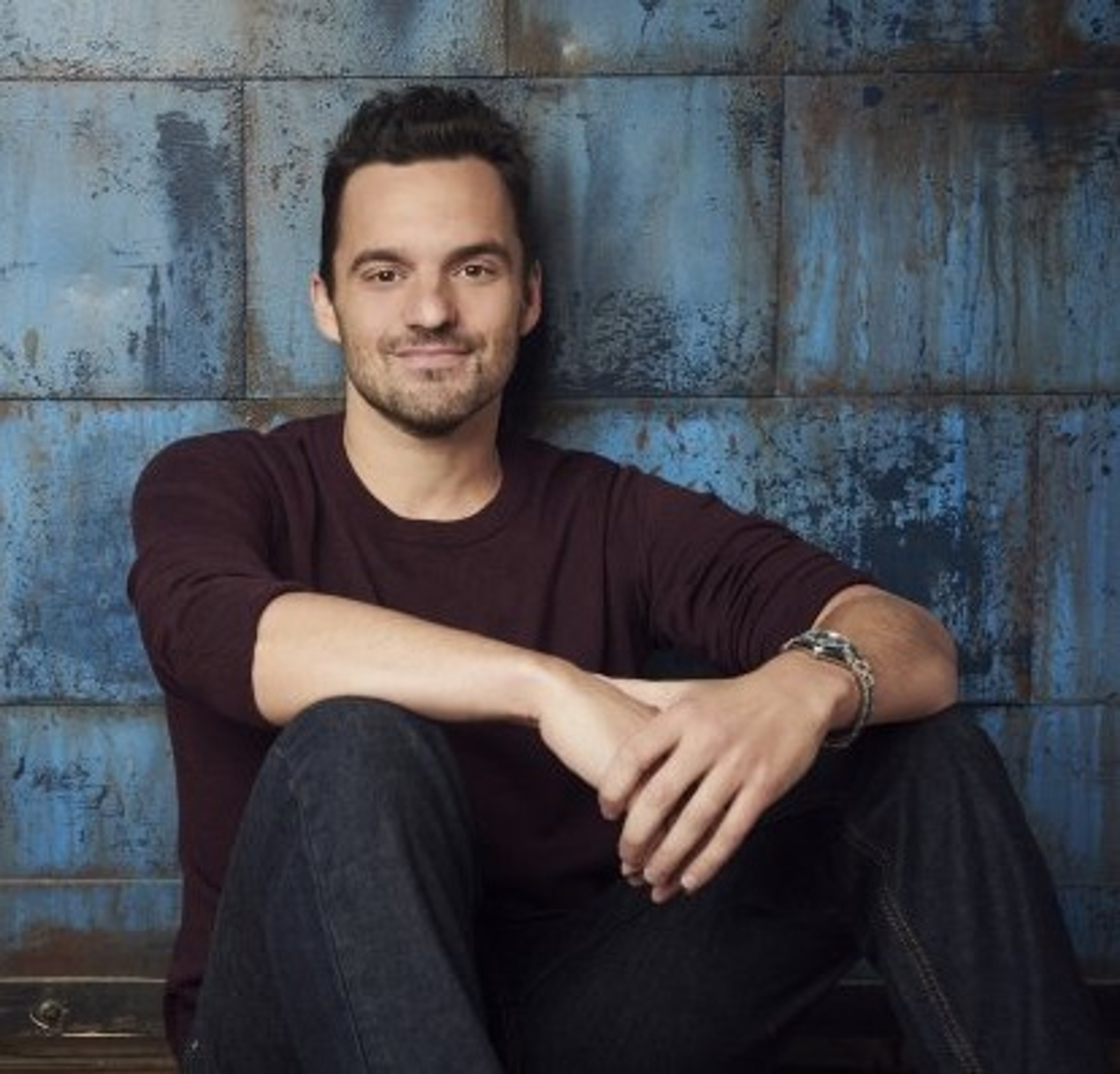 A man with dark hair and a beard, wearing a burgundy shirt, sitting against a distressed blue wall.