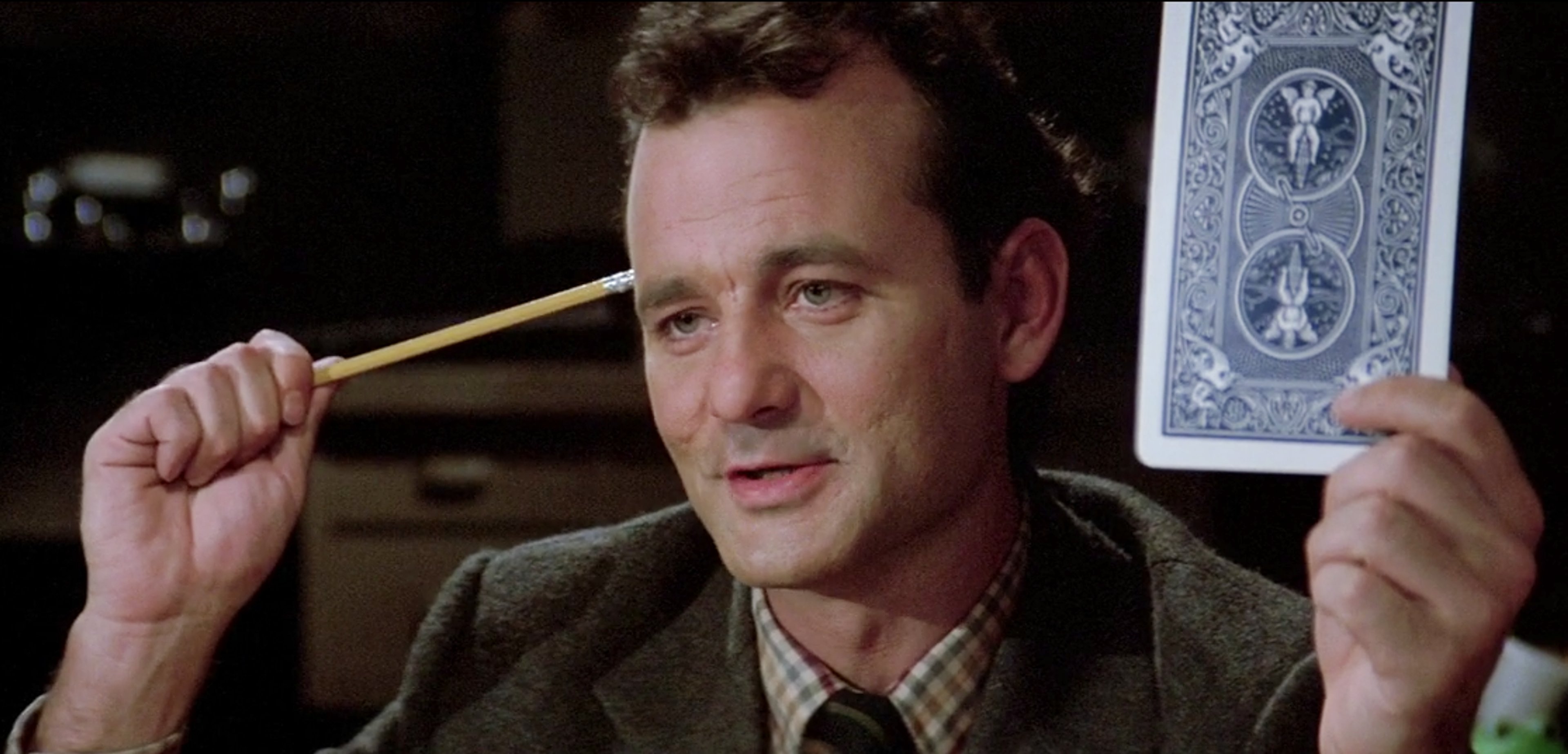 A man with curly brown hair and a mustache wearing a tan jacket and tie, holding a card or document and looking slightly sarcastic.