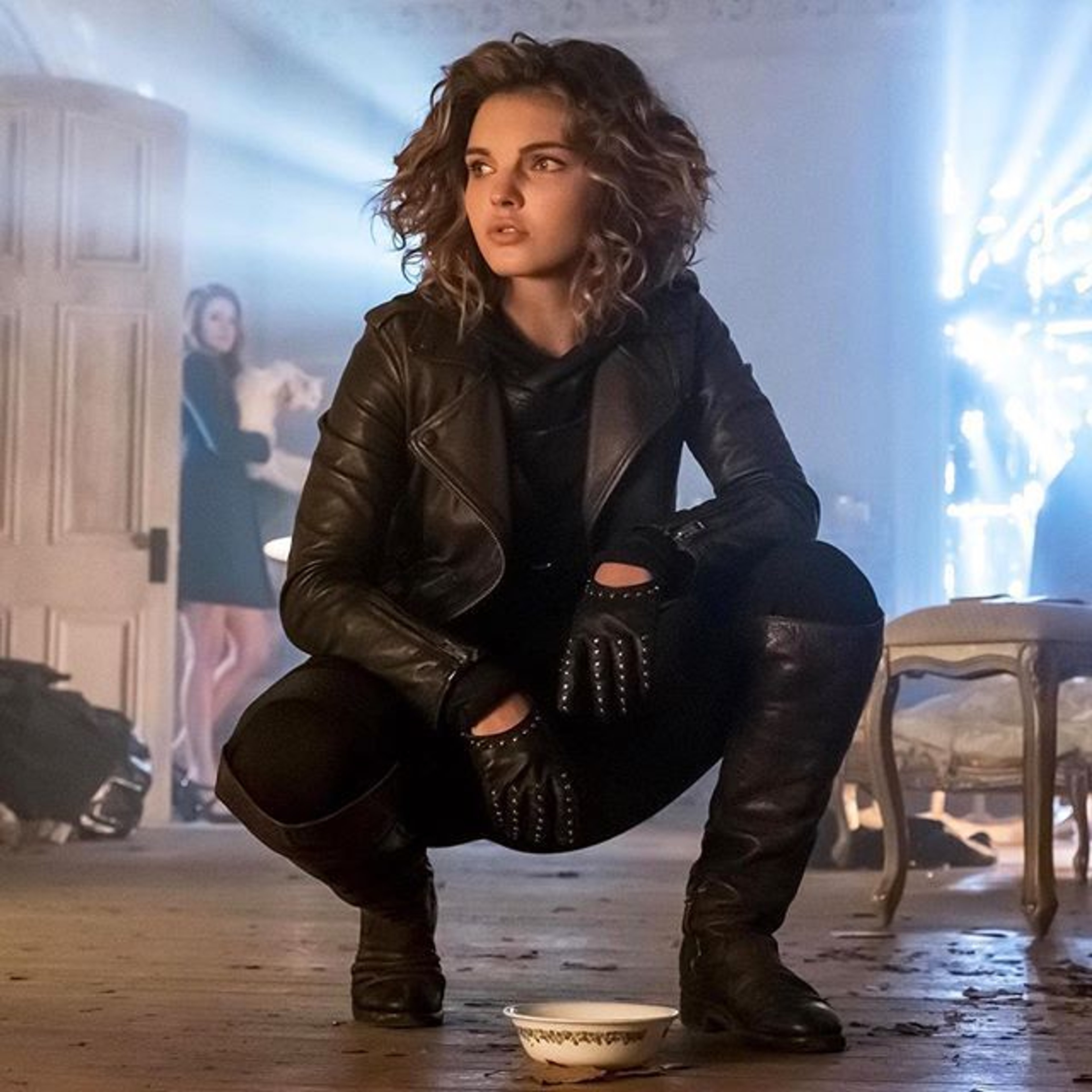 A young woman with curly dark hair wearing a black leather jacket and tight black pants crouching in an urban setting.