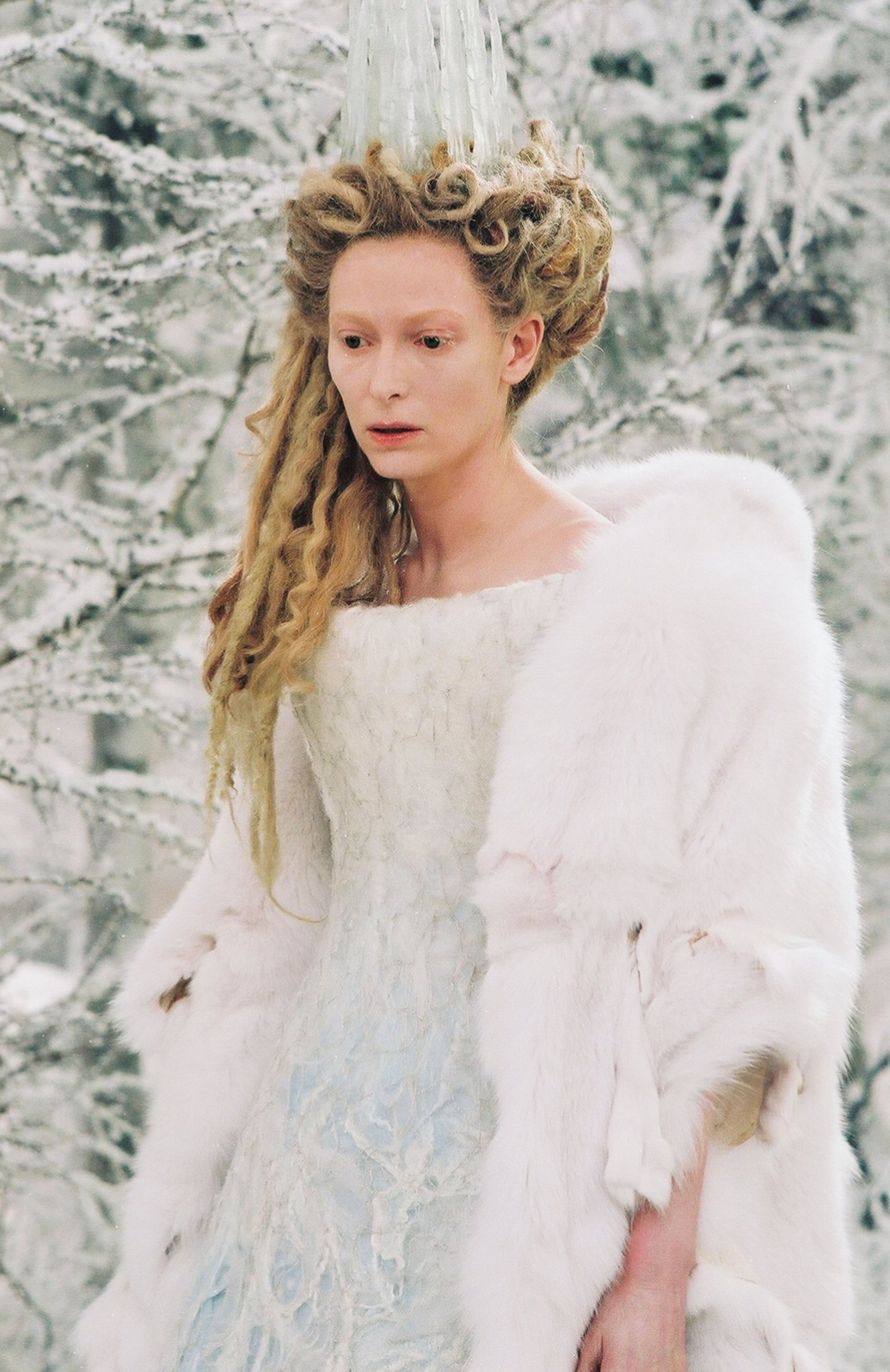 A woman with long, curly blonde hair wearing a white fur-trimmed cloak, appearing to be the character Jadis, the White Witch, from the Chronicles of Narnia.