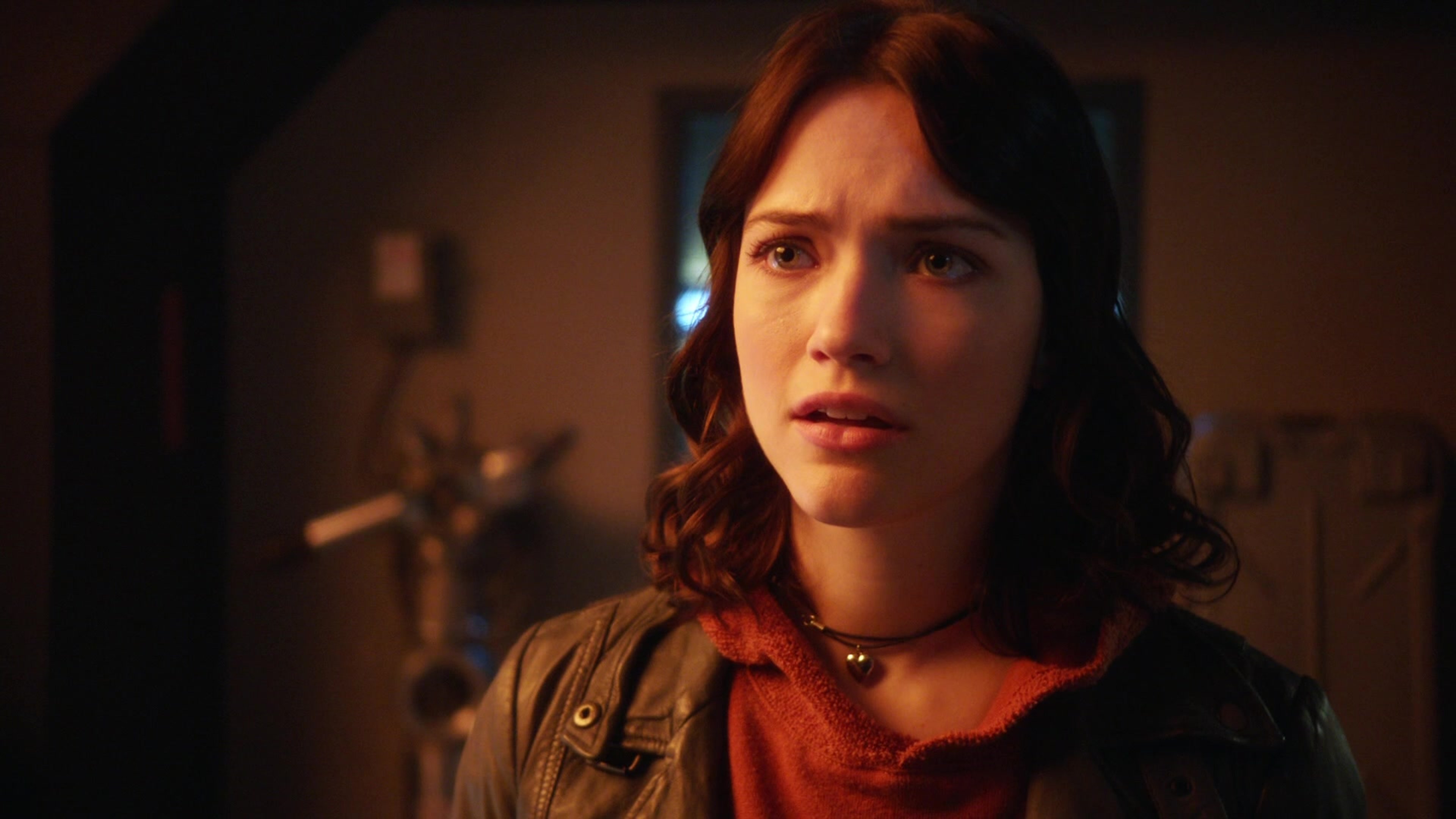 A young woman with long brown hair wearing a red jacket in a dimly lit setting.