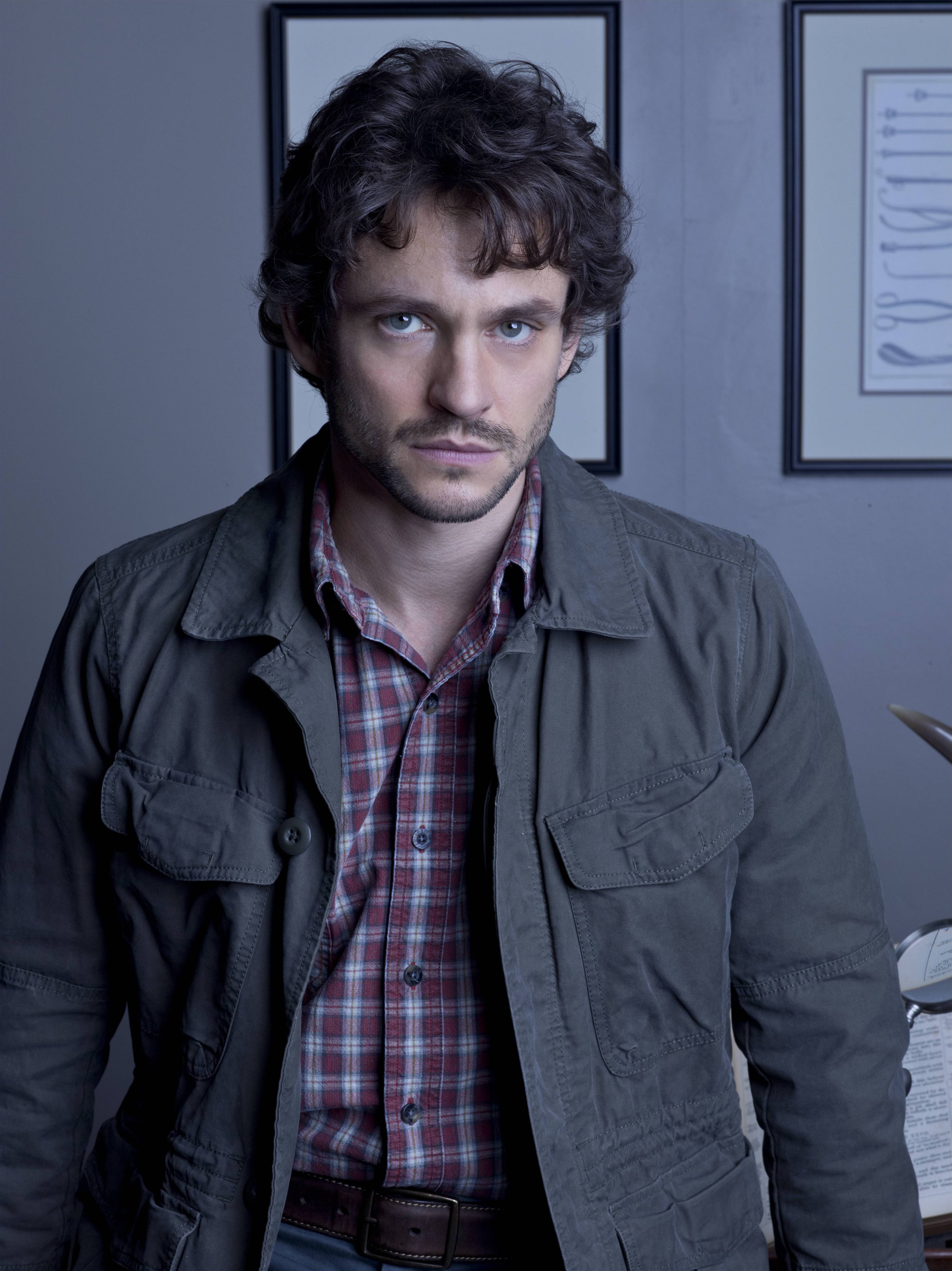 A man with curly dark hair wearing a plaid shirt and gray jacket