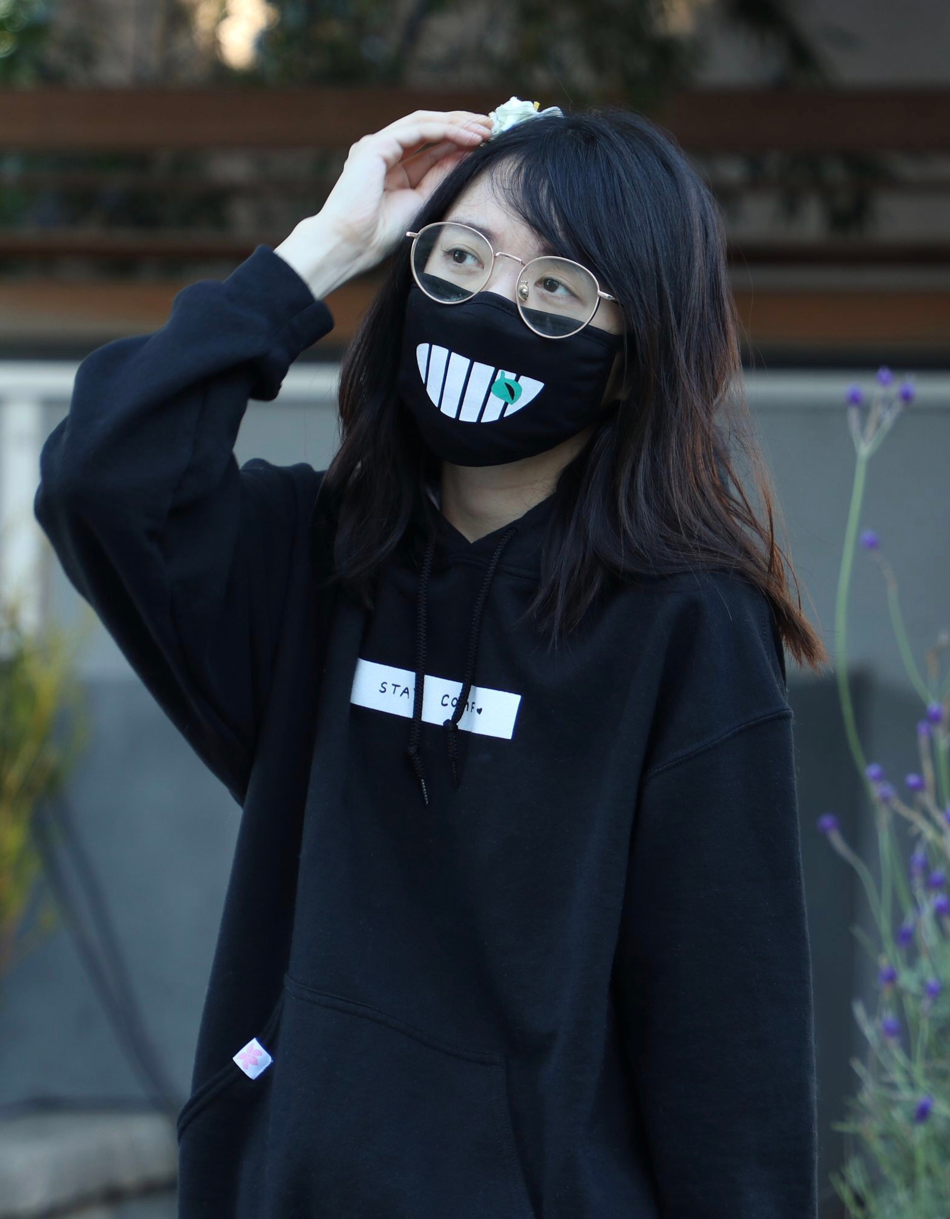 A young woman wearing a black hoodie and a mask with a large grinning mouth design, standing outdoors.