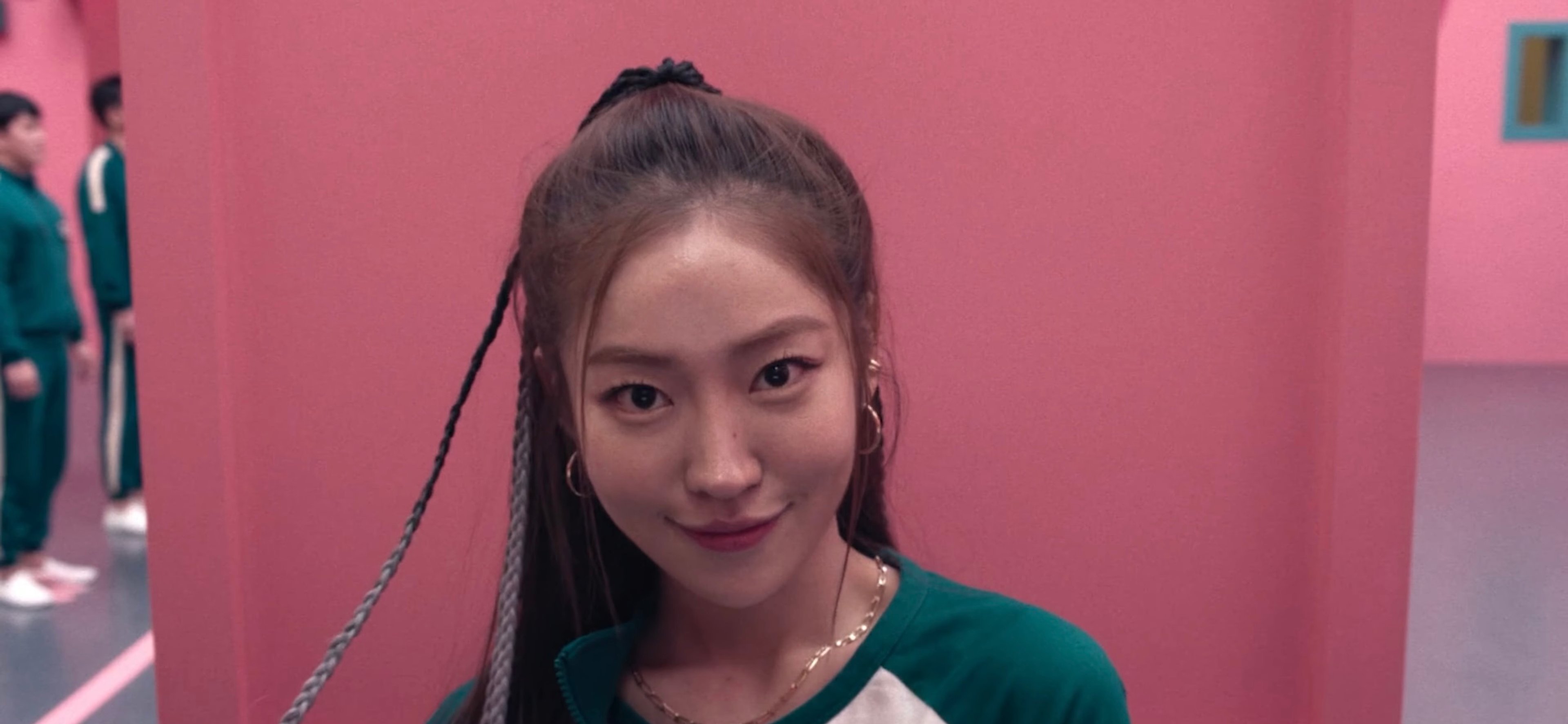 A young woman with long dark hair in a green sweater smiling at the camera against a pink and blue background.