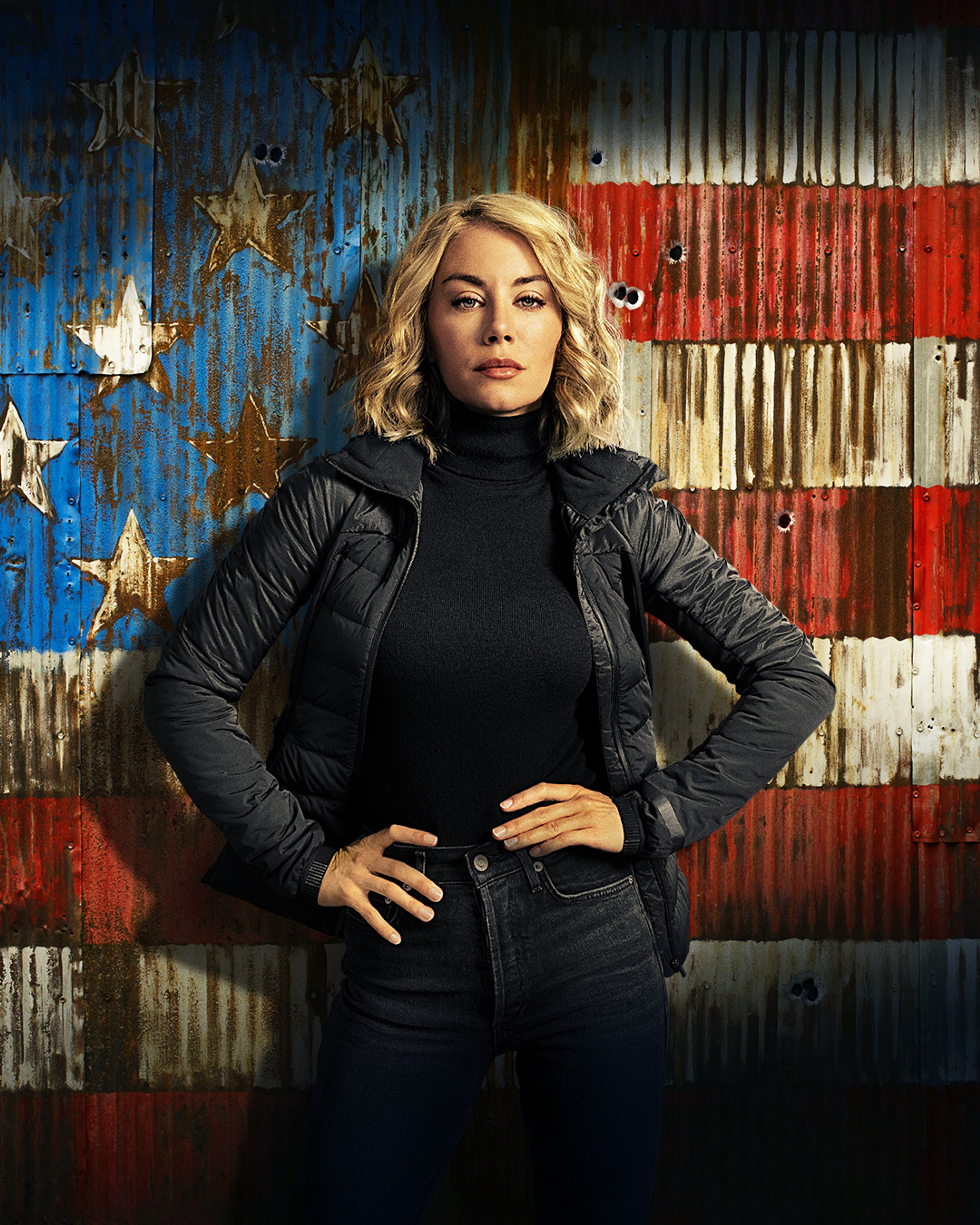 A woman with blonde hair wearing a black turtleneck sweater standing in front of an American flag backdrop.
