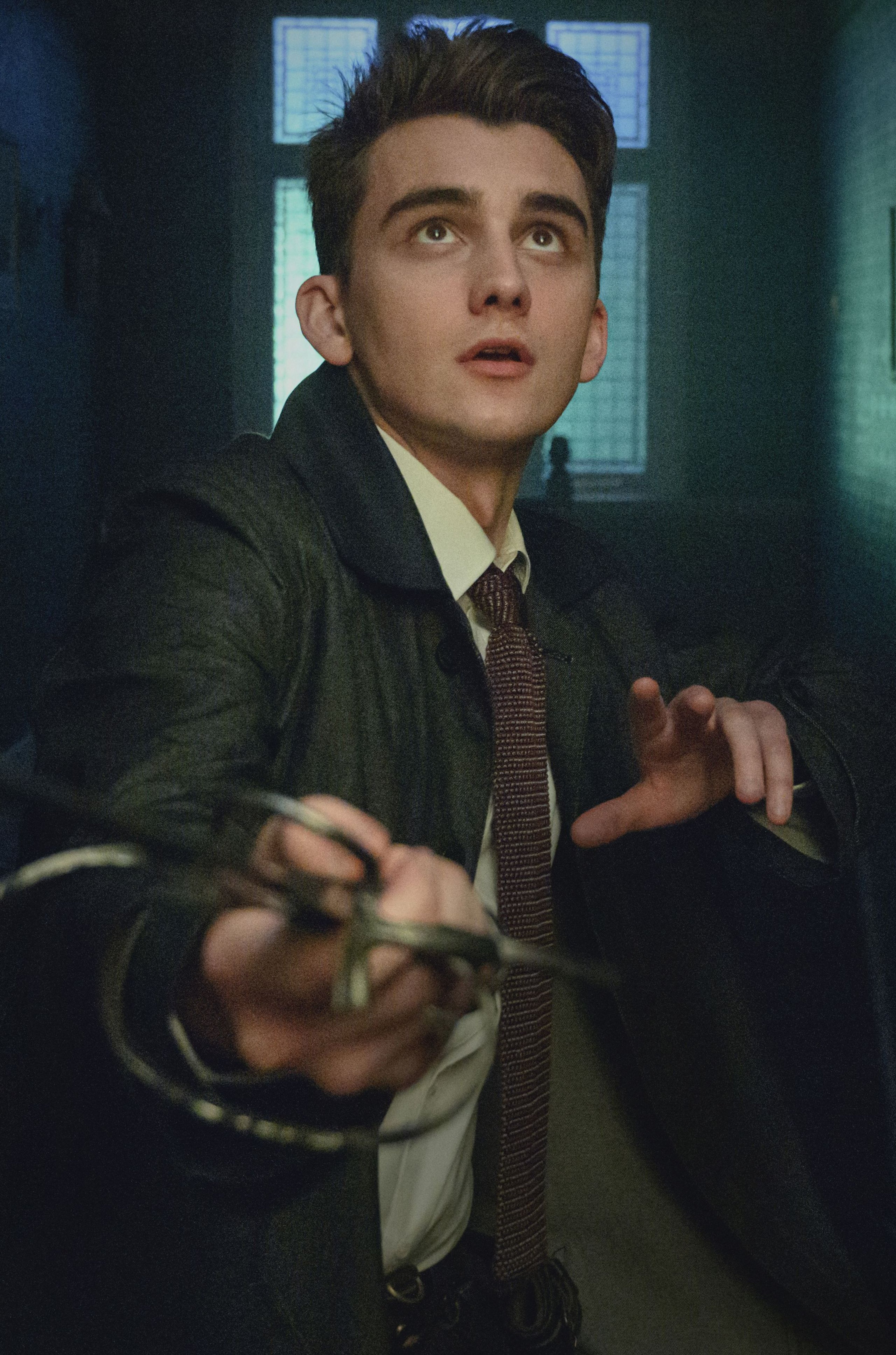 A young man with dark hair wearing a green coat and tie, holding an object.