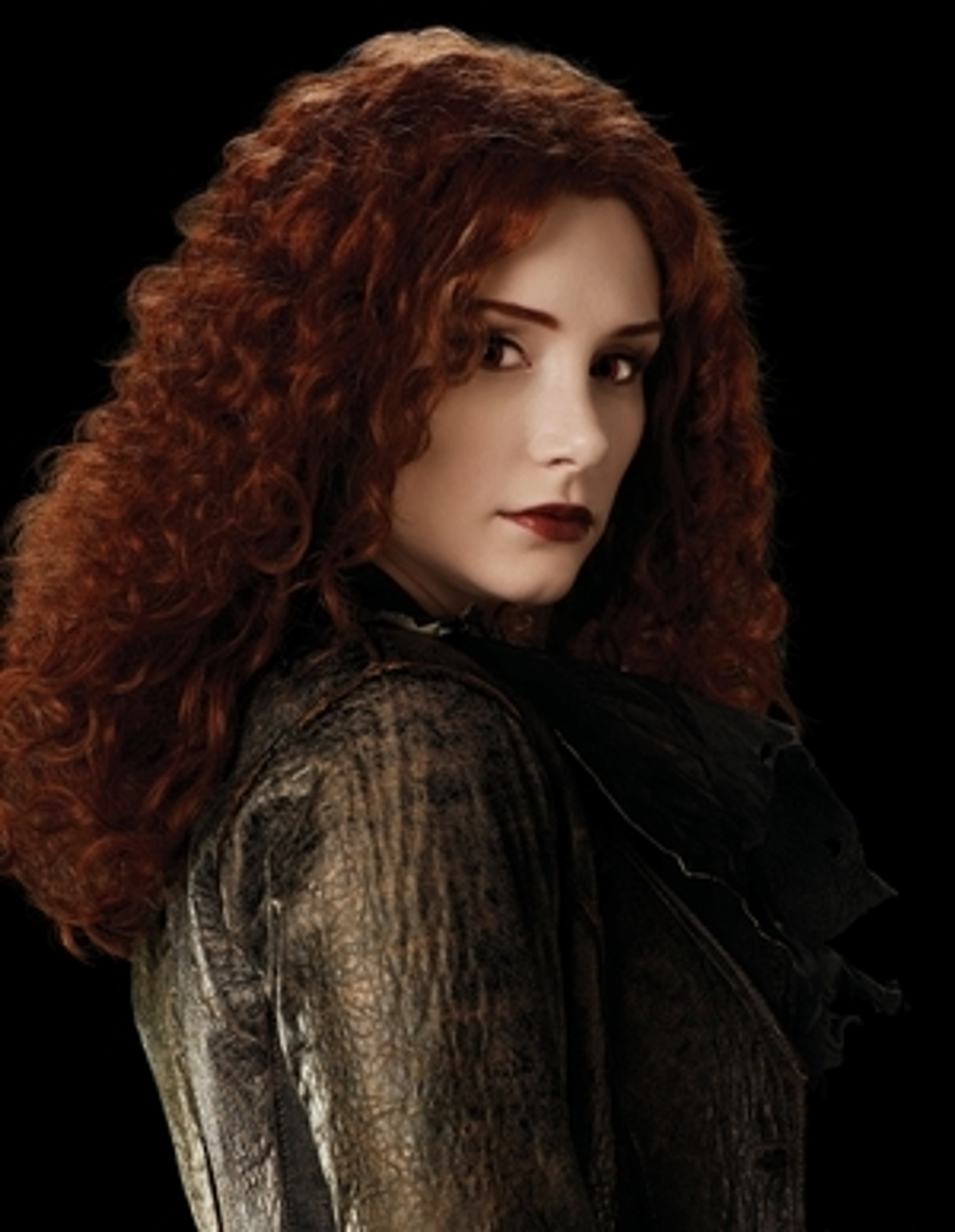 A woman with long, curly red hair wearing a dark dress stands in a dramatic, shadowy setting.