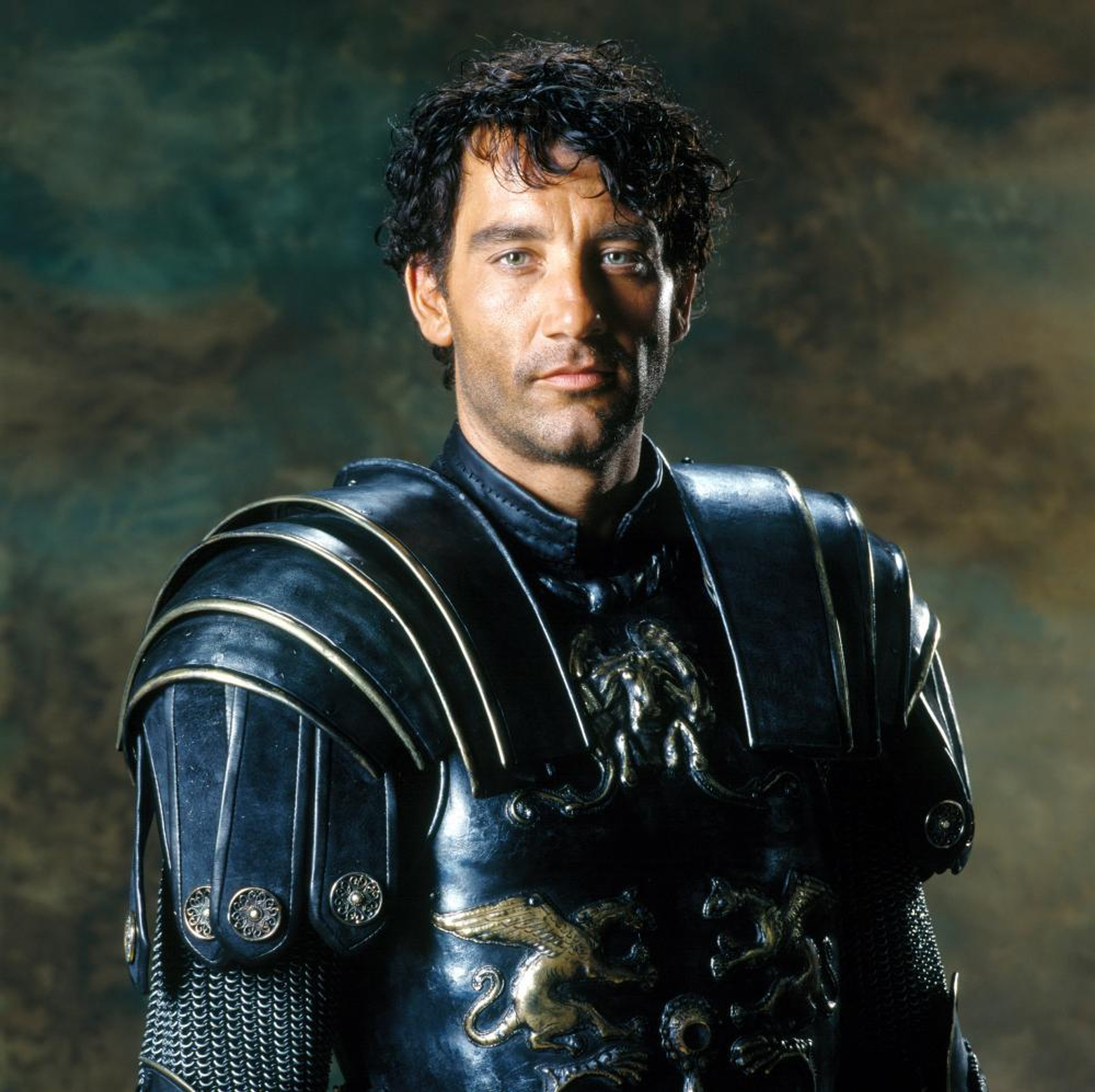 A man with dark curly hair wearing a black leather outfit with armor-like elements, looking directly at the camera with an intense gaze.