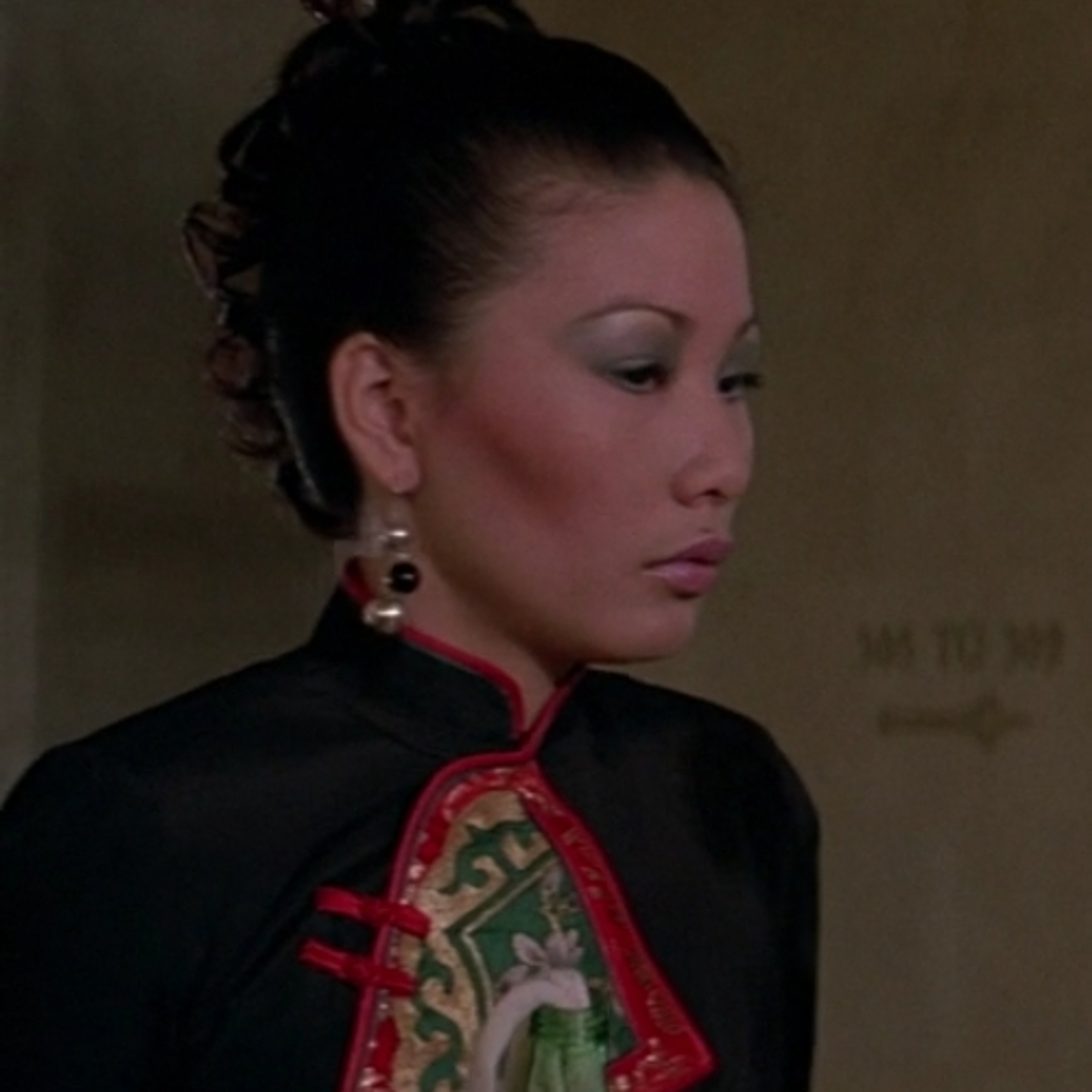 A woman with dark hair in an elegant updo, wearing a red and black outfit with a high collar, in a formal or professional setting.